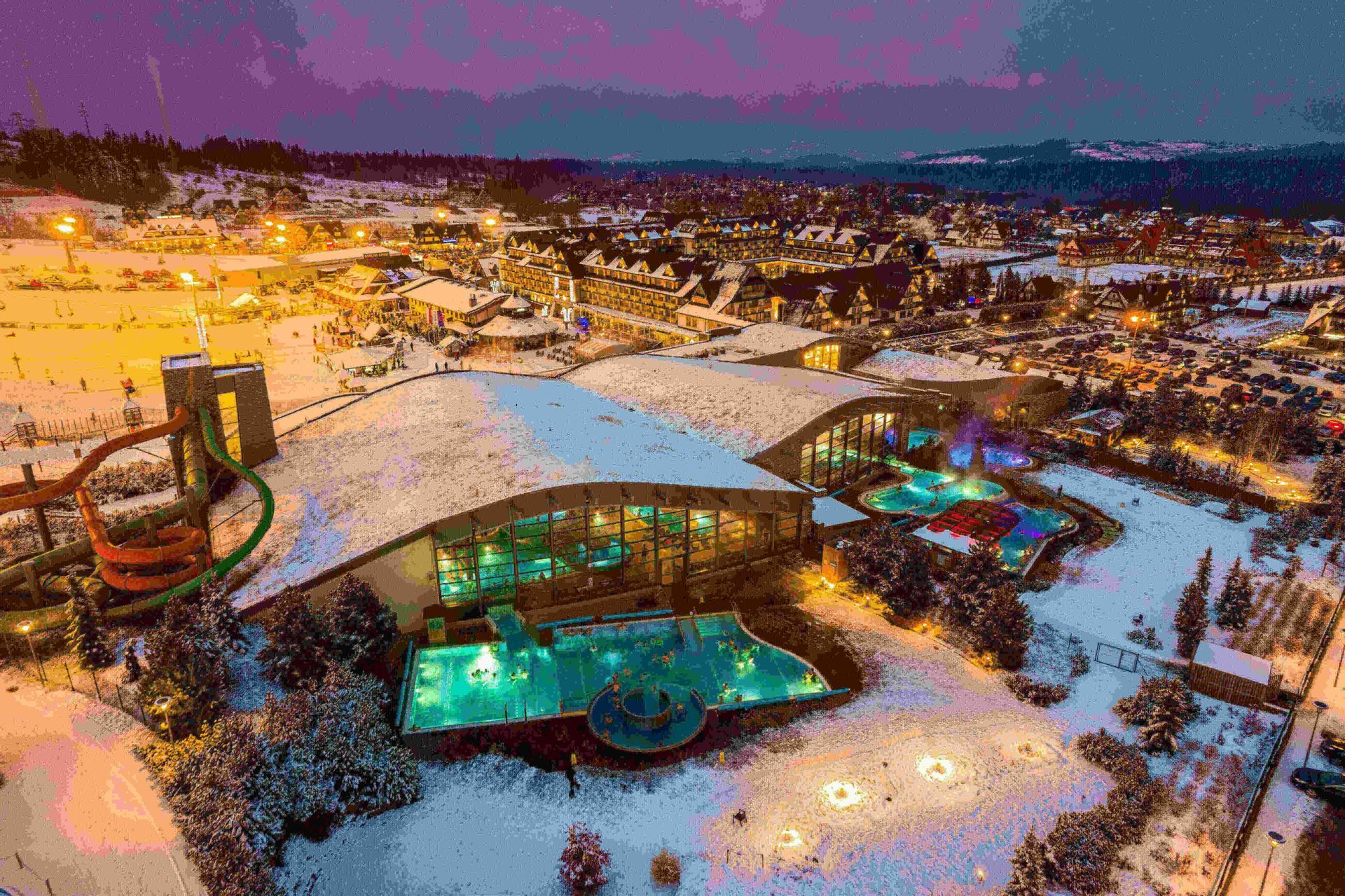 Una vista aérea de un gran complejo de spa con piscinas turquesa iluminadas, situado en un pueblo cubierto de nieve al atardecer.