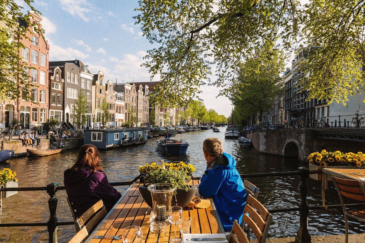 Deux personnes assises à une table en bois sur une terrasse, regardant un canal de la ville avec des bateaux et des bâtiments.