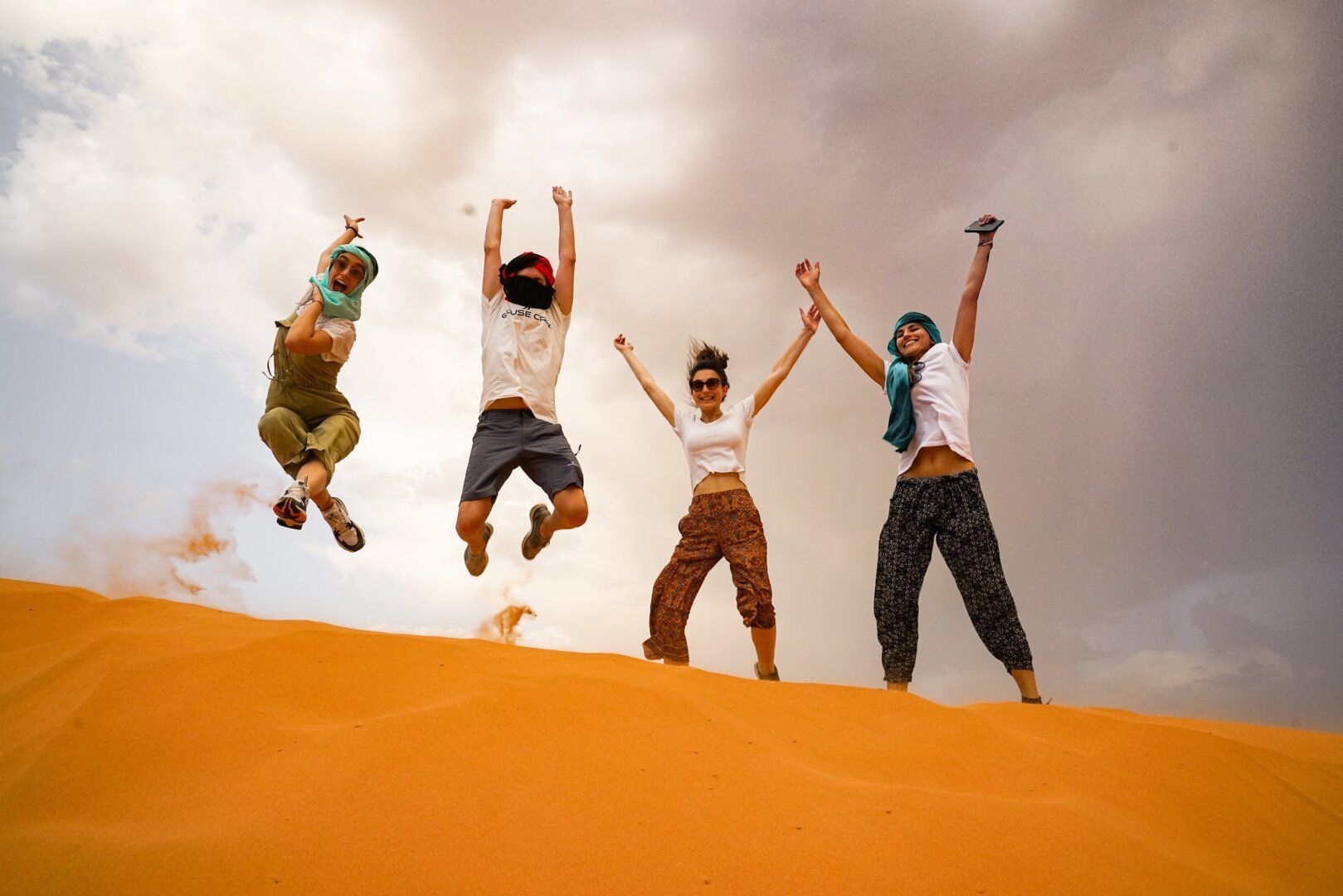 Un voyage de groupe WeRoad de quatre personnes sautant en l'air sur une dune de sable orange sous un ciel partiellement nuageux.