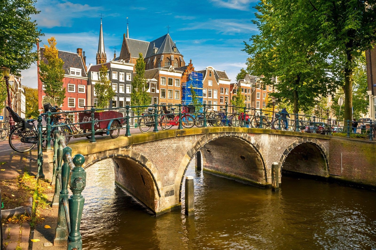 Un pont en arc de brique avec des vélos garés traverse un canal de ville, avec des maisons à pignons traditionnelles et des arbres le long de la berge.