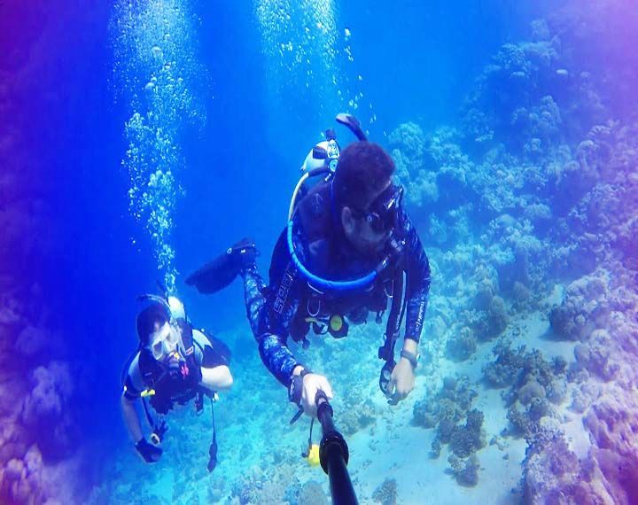 Deux personnes d'un voyage de groupe WeRoad faisant de la plongée sous-marine, l'une prenant une photo avec une perche à selfie au-dessus d'un récif corallien.