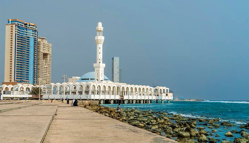 Une mosquée blanche avec un minaret et un dôme bleu se dresse sur une jetée au-dessus de la mer, avec un littoral rocheux et des bâtiments urbains modernes en arrière-plan.