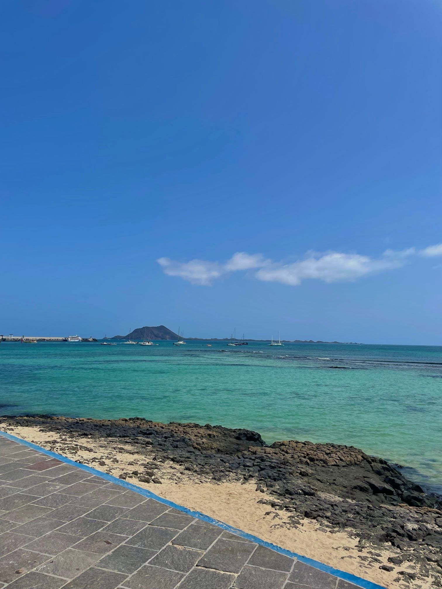 Un paseo de piedra con vistas a una bahía de aguas turquesas, donde veleros están anclados con una gran isla al fondo bajo un cielo azul.