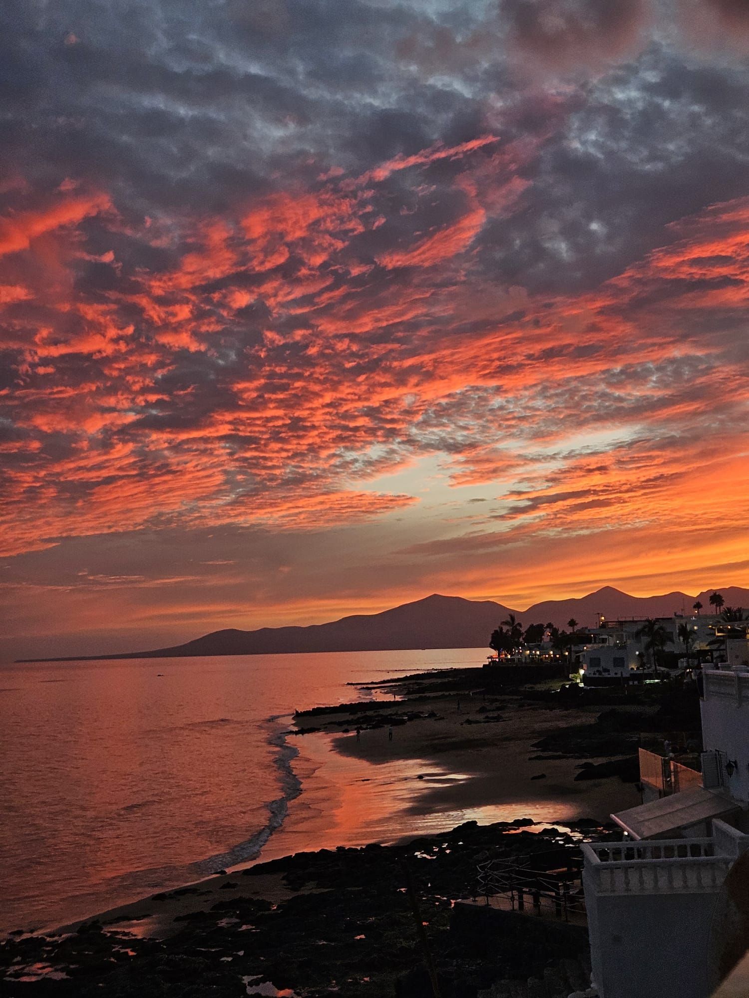 Nubes dispersas rojas y naranjas durante el atardecer sobre el océano, con una costa y montañas silueteadas al fondo.