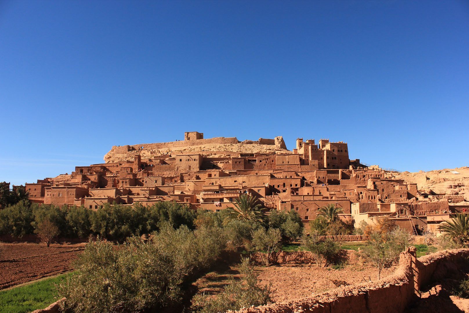 A traditional mud-brick village built on a hillside under a clear blue sky, with trees and fields in the foreground.