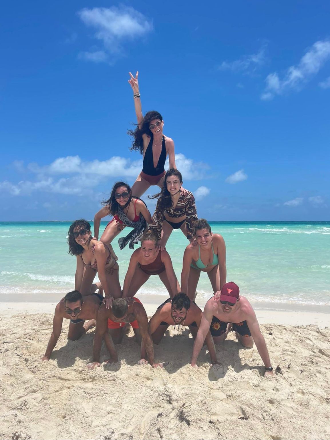 Un viaje en grupo de WeRoad forma una pirámide humana en una playa de arena frente a un mar turquesa bajo un cielo azul claro.