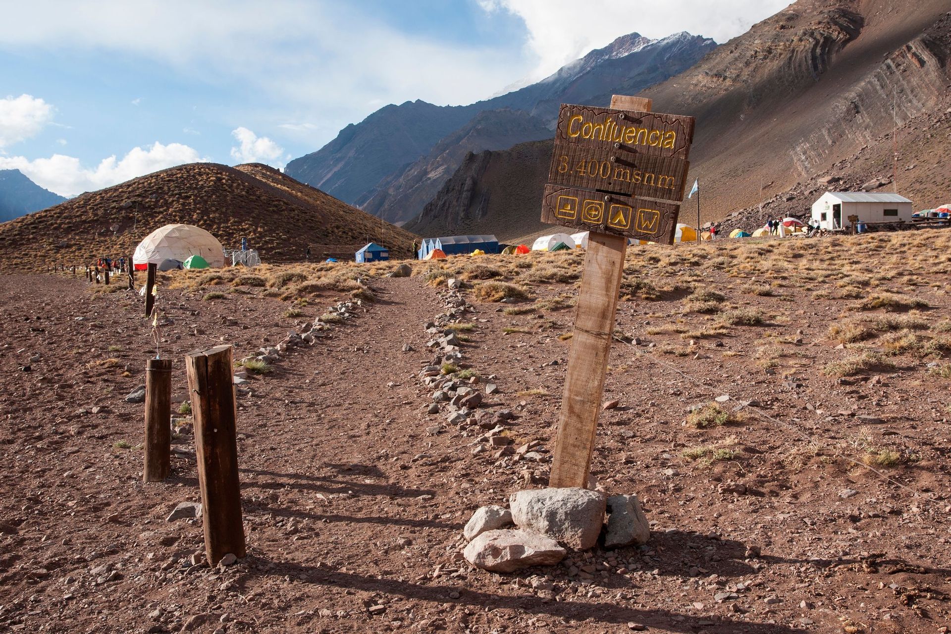 A wooden sign for Confluencia camp sits on a rocky path leading to a high-altitude campsite with tents at the base of mountains.