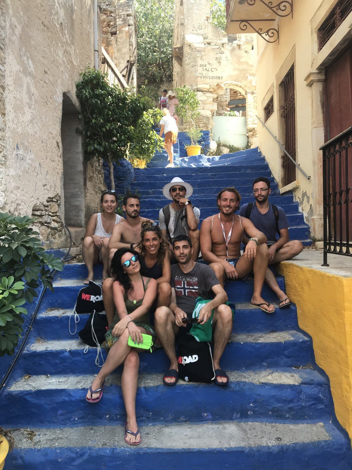 Eine WeRoad-Reisegruppe von acht Personen sitzt gemeinsam auf einer breiten, blau gestrichenen Treppe in einer engen Dorfgasse.