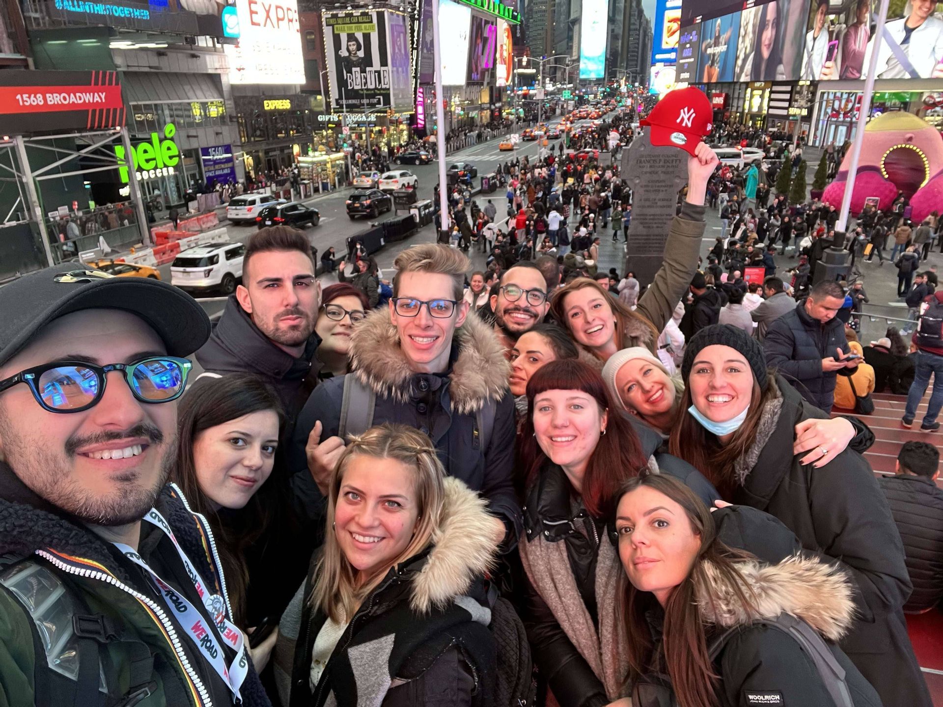 Un grupo de WeRoad se toma una selfie en unas escaleras con vistas a una animada plaza de la ciudad, llena de luminosos carteles y multitudes.
