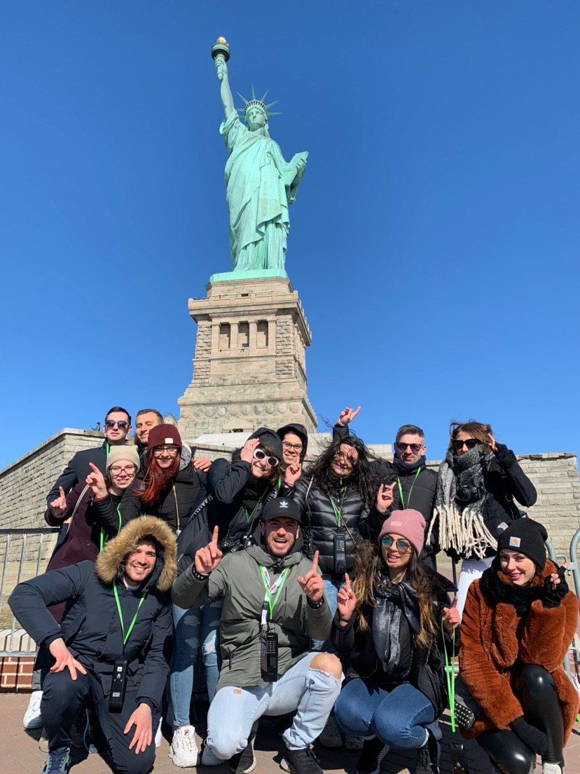 Un gruppo WeRoad in posa per una foto davanti alla Statua della Libertà sotto un cielo azzurro e limpido.