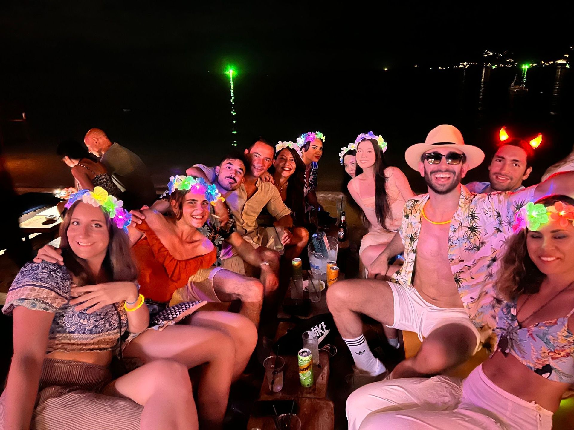 Un gruppo WeRoad posa per una foto a una festa in spiaggia notturna, indossando cerchietti floreali luminosi e festosi.