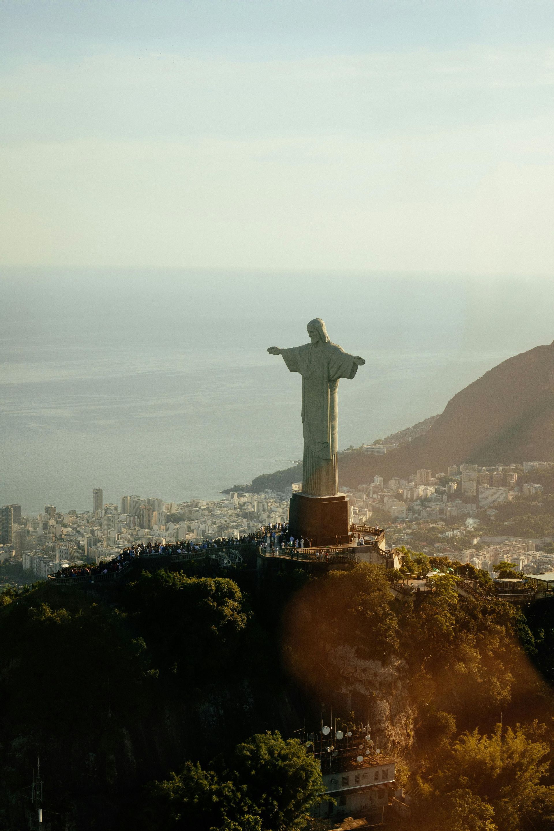 A large statue with outstretched arms stands on a verdant mountain peak, overlooking a coastal city with a crowd of people at its base.