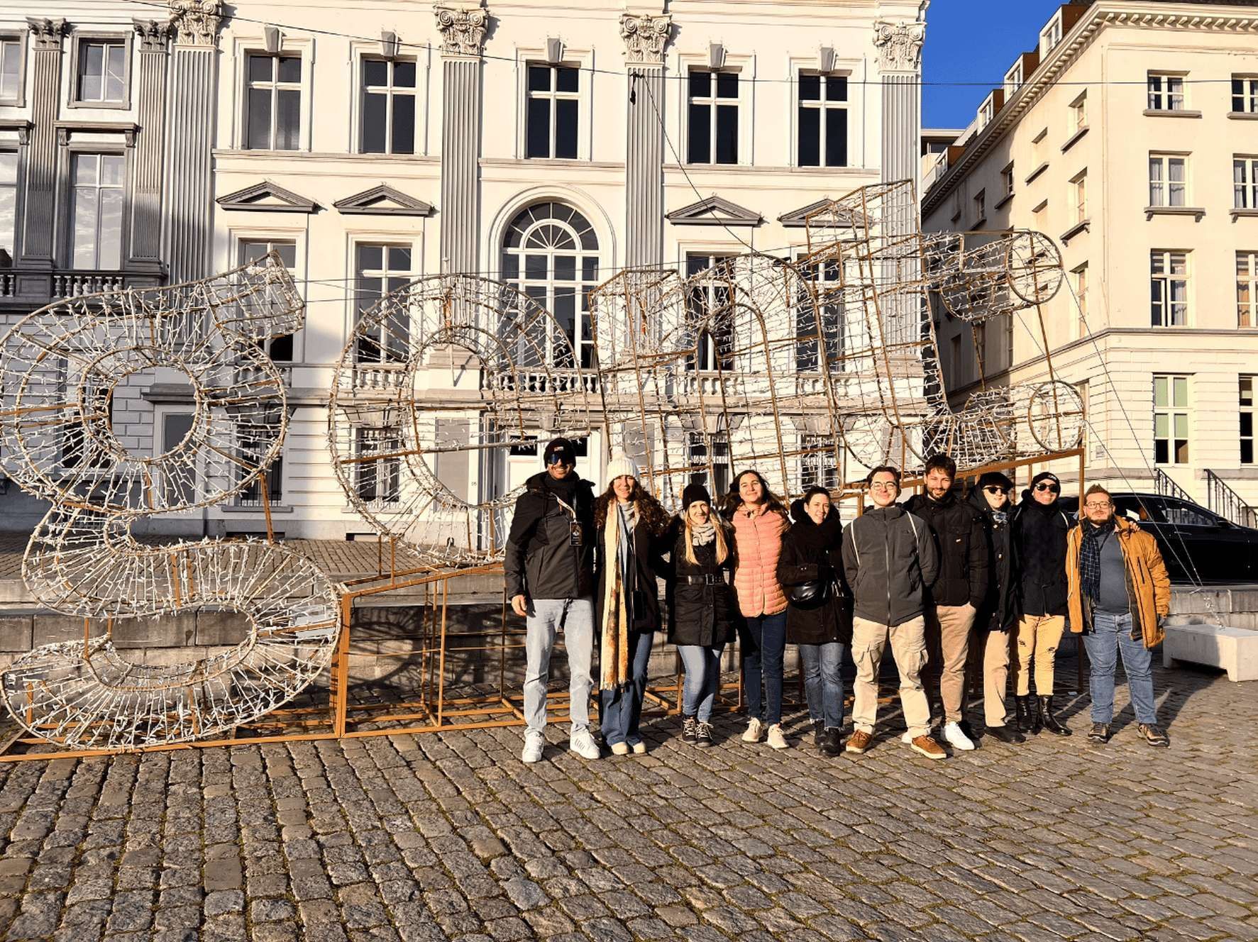 Un gruppo WeRoad in posa su una strada acciottolata davanti a una grande installazione luminosa e un edificio classico.
