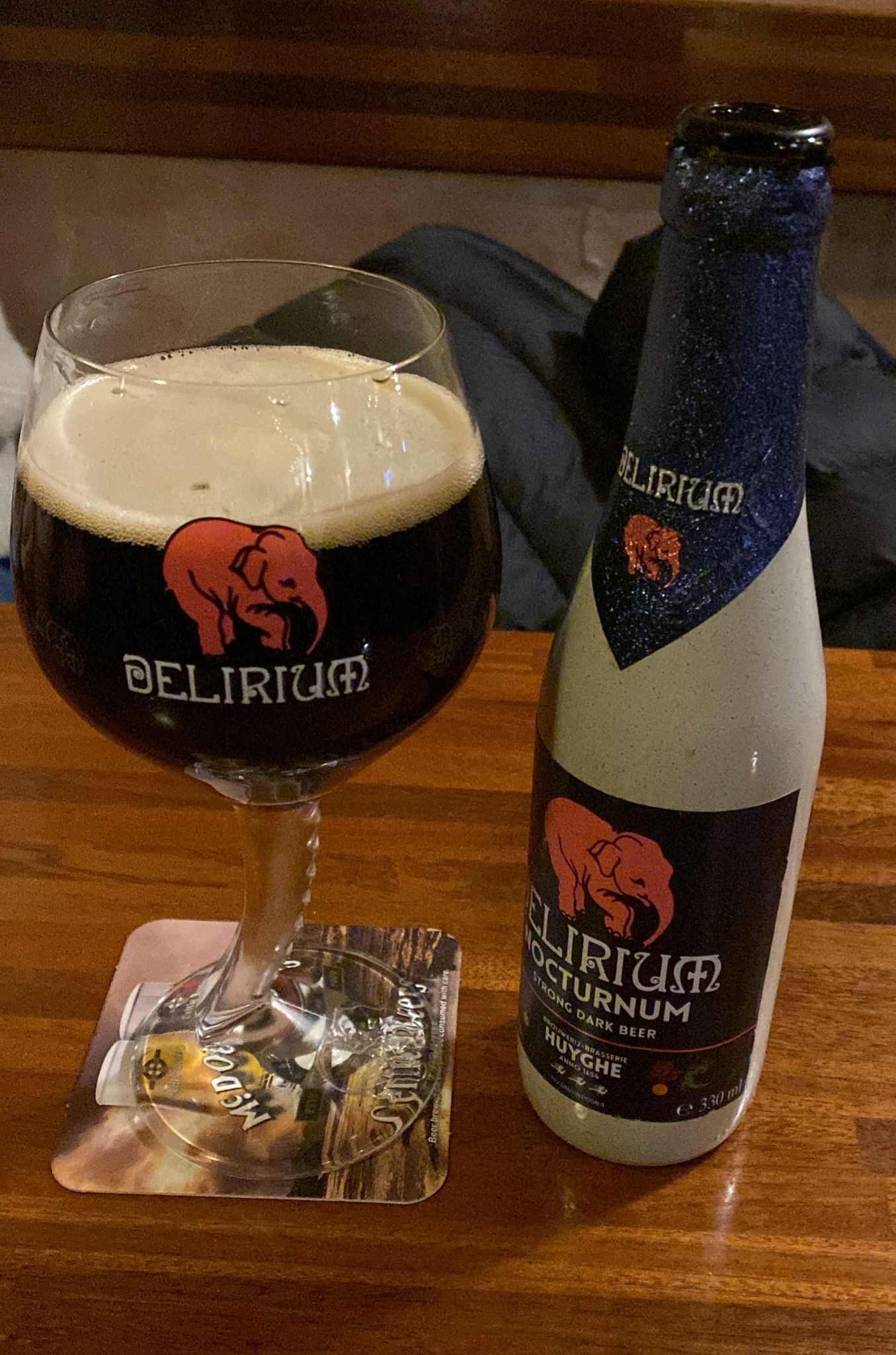Un bicchiere di birra scura Delirium Nocturnum con una schiuma densa, posto accanto alla sua bottiglia abbinata su un tavolo di legno.