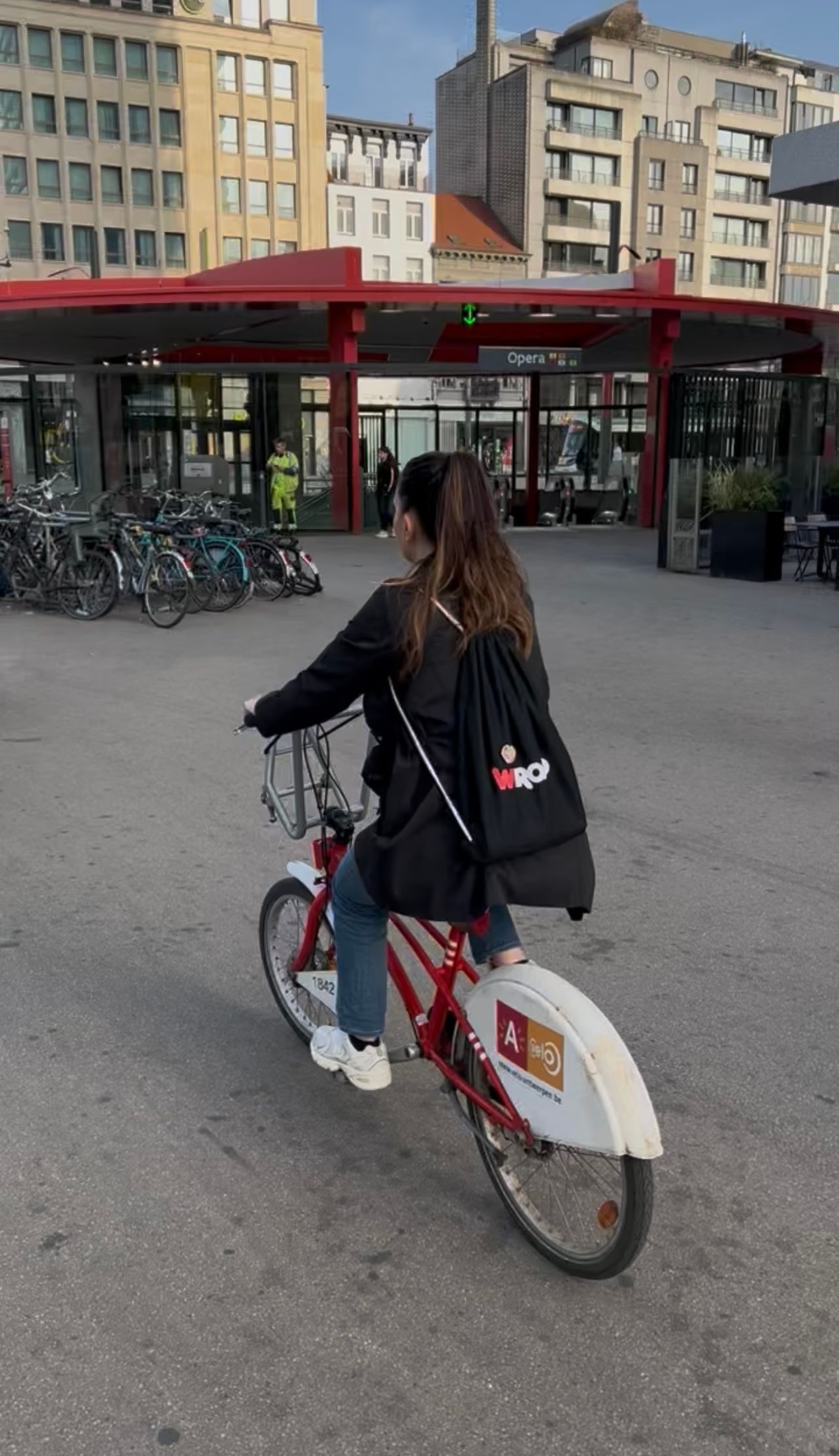 Una donna vista di schiena, con uno zaino WeRoad nero, pedala su una bicicletta rossa attraverso una piazza cittadina pavimentata.