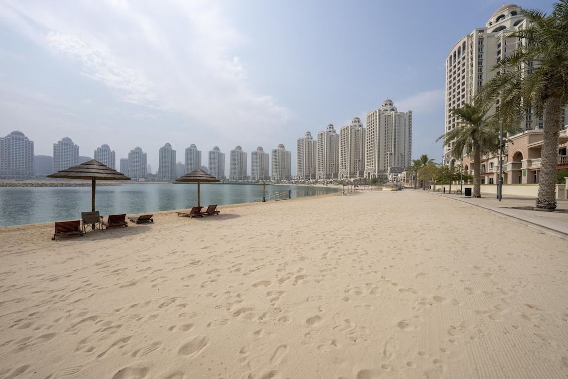 Un'ampia spiaggia sabbiosa con ombrelloni e sedie a sdraio di fronte a un moderno skyline cittadino oltre l'acqua.