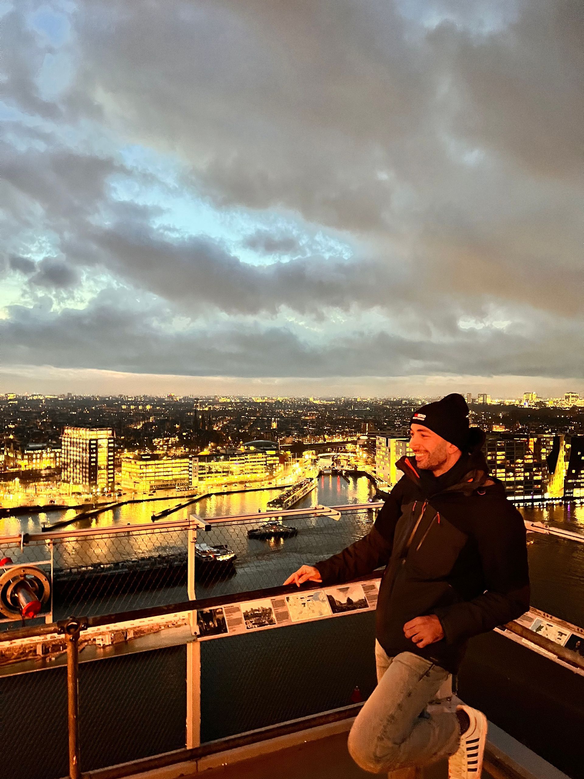 Un uomo con berretto e giacca si trova su una piattaforma panoramica, che si affaccia su un paesaggio urbano illuminato e un fiume al tramonto.