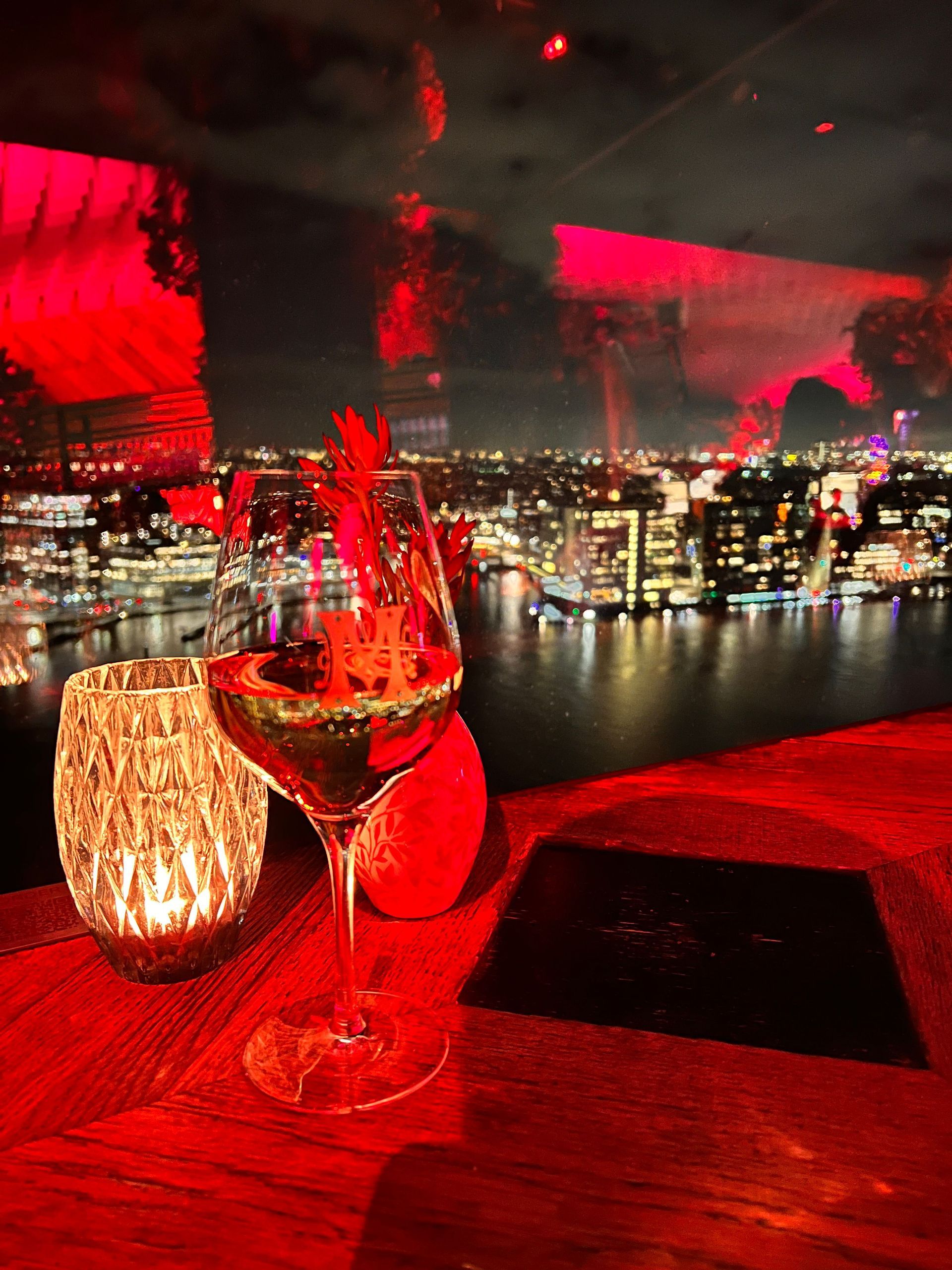 Un bicchiere di vino e una candela su un tavolo in luce rossa, con vista su uno skyline illuminato della città oltre un fiume di notte.