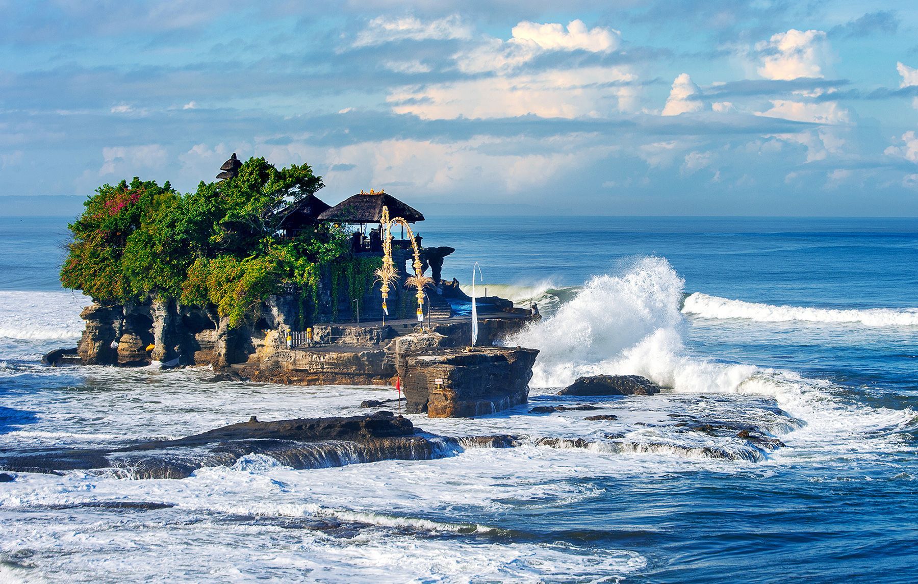 Un templo se asienta en una pequeña isla rocosa cubierta de árboles, con grandes olas del océano rompiendo contra la orilla bajo un cielo parcialmente nublado.