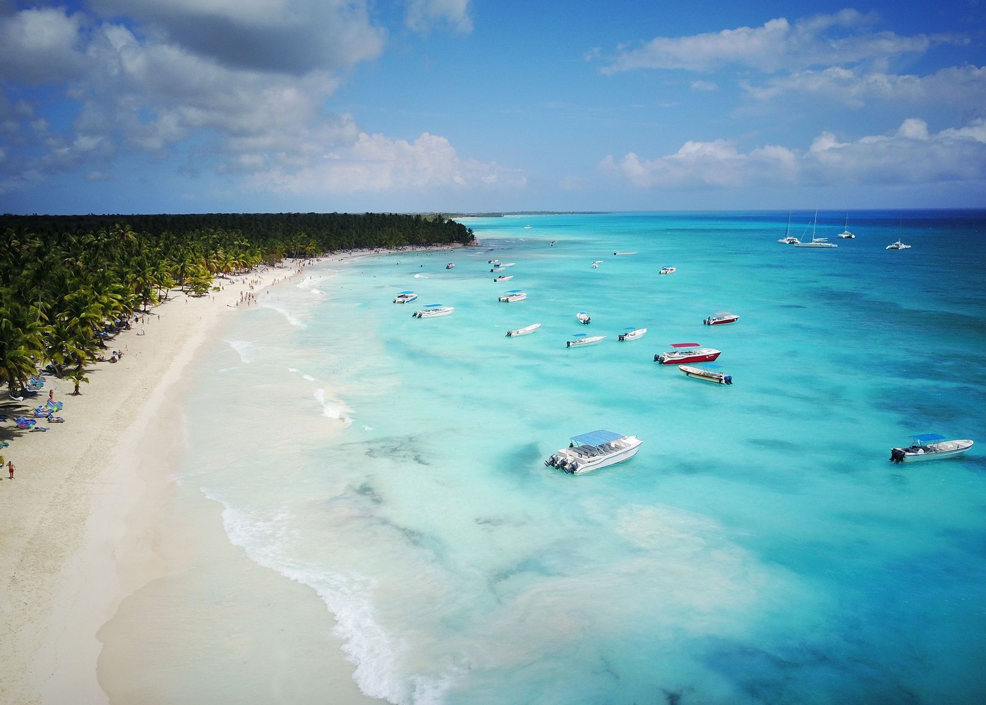 Una vista aerea di una spiaggia tropicale con sabbia bianca, palme e numerose barche ancorate in acque limpide e turchesi.