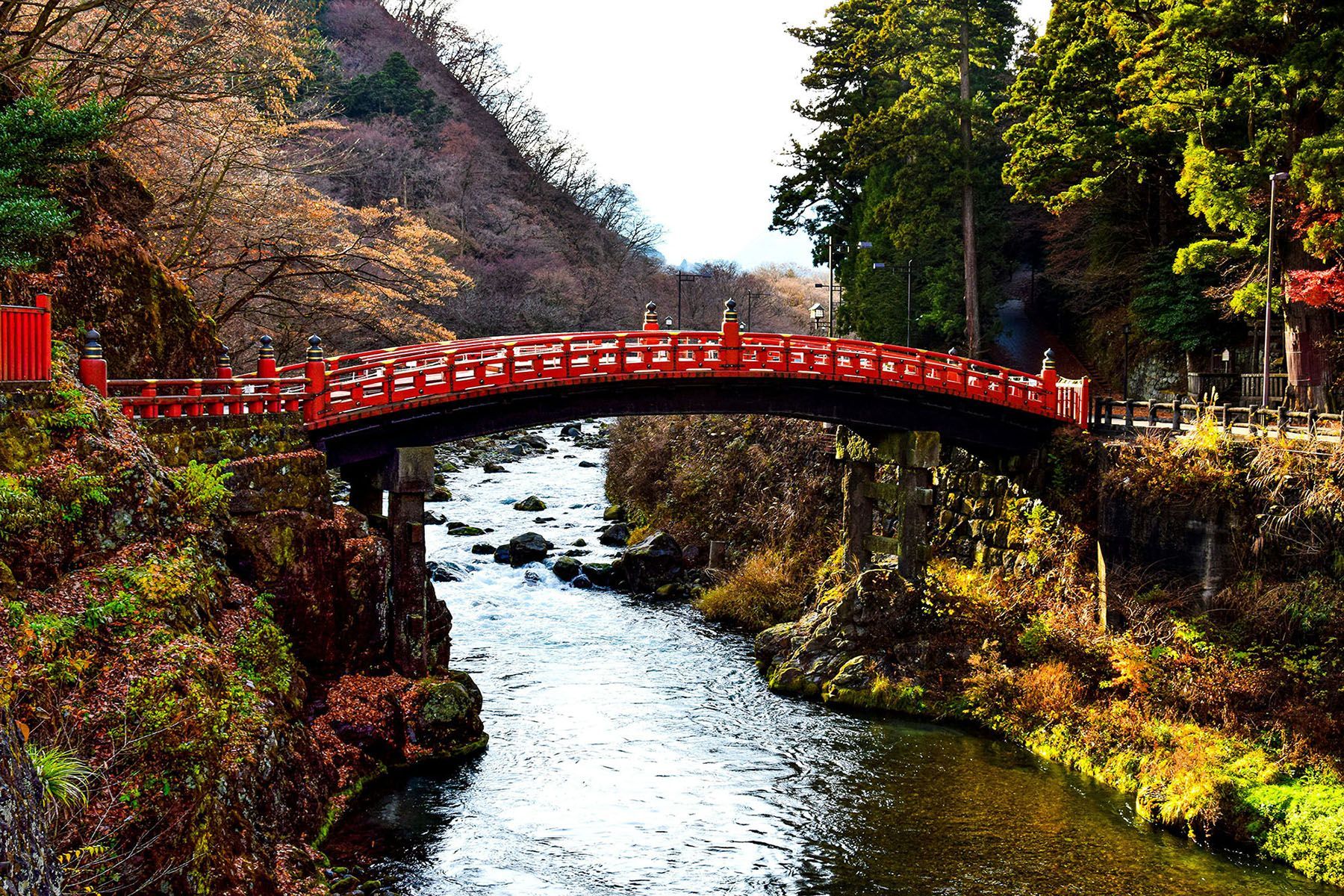 Un puente rojo y ornamentado con arcos cruza un río rocoso que fluye por un valle boscoso con follaje de otoño.