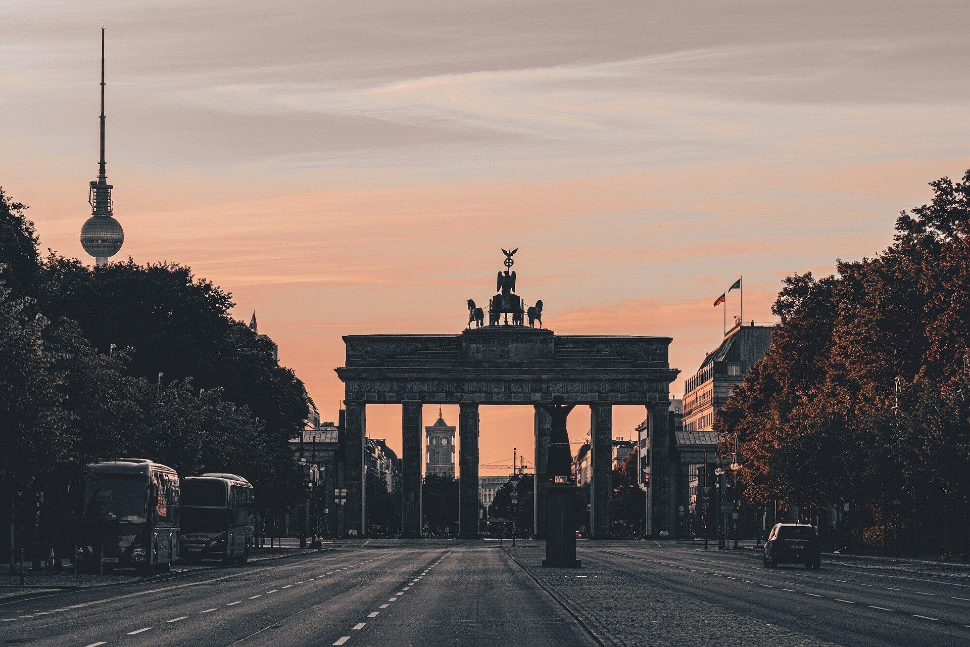 La Puerta de Brandeburgo se alza silueteada al final de una calle ancha contra un cielo anaranjado al atardecer, con la Torre de Televisión de Berlín al fondo.