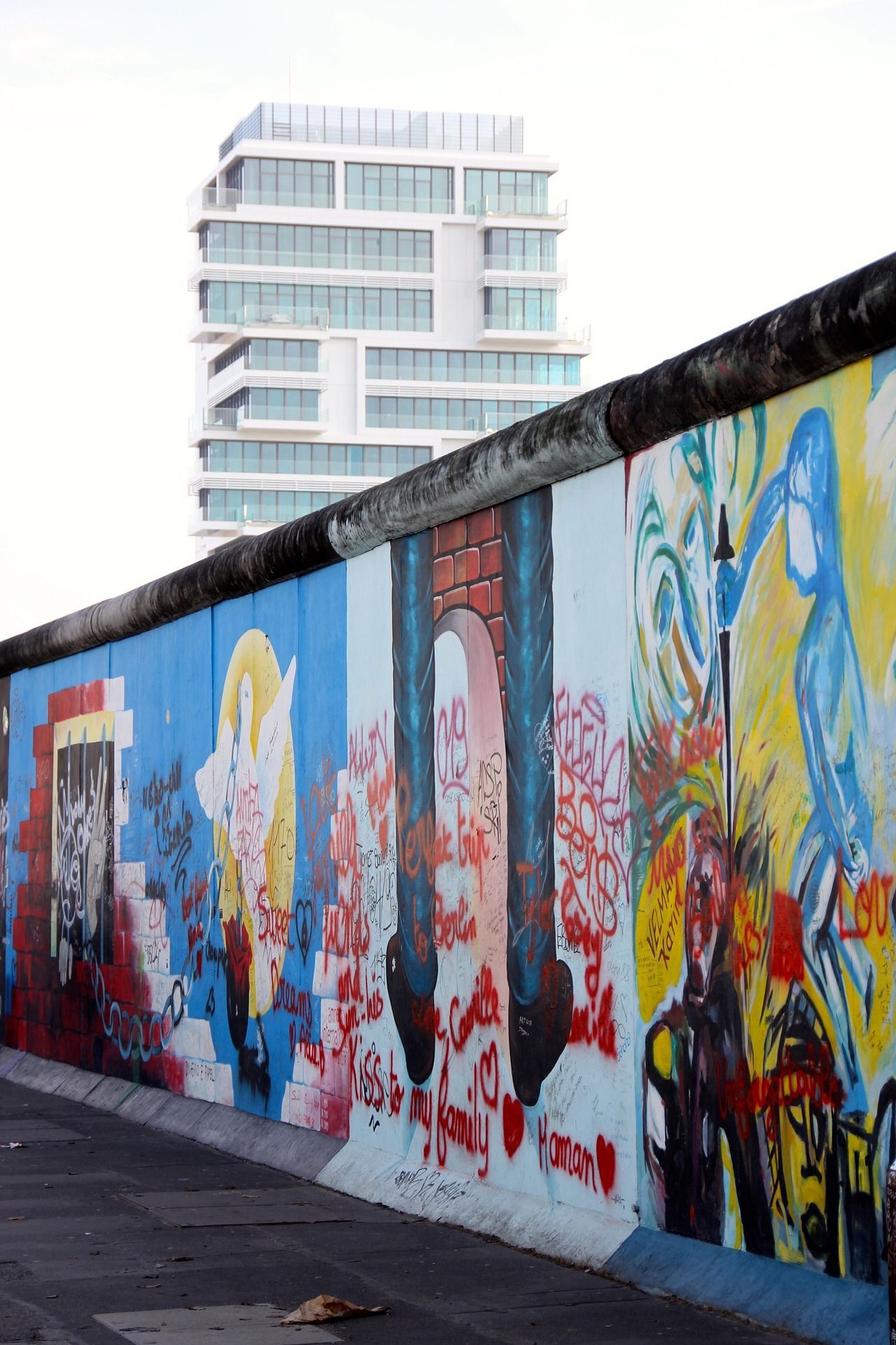 Una larga pared de hormigón cubierta de coloridos murales y grafitis, con un edificio moderno visible al fondo.