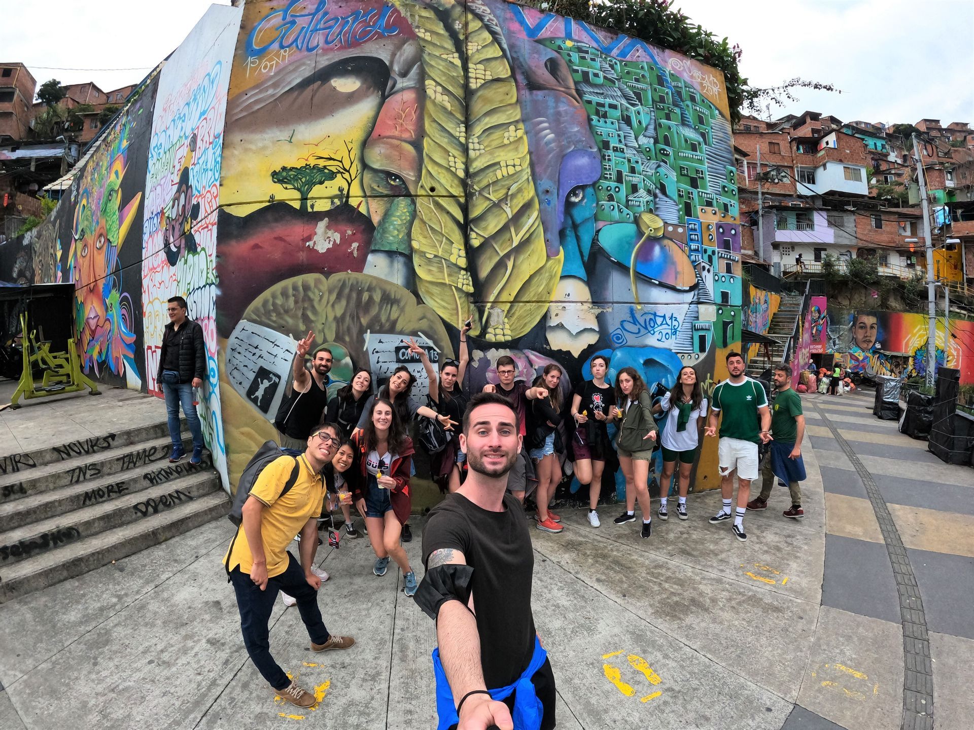 Un groupe WeRoad en voyage pose pour un selfie devant une grande fresque de graffiti colorée dans un quartier à flanc de colline.