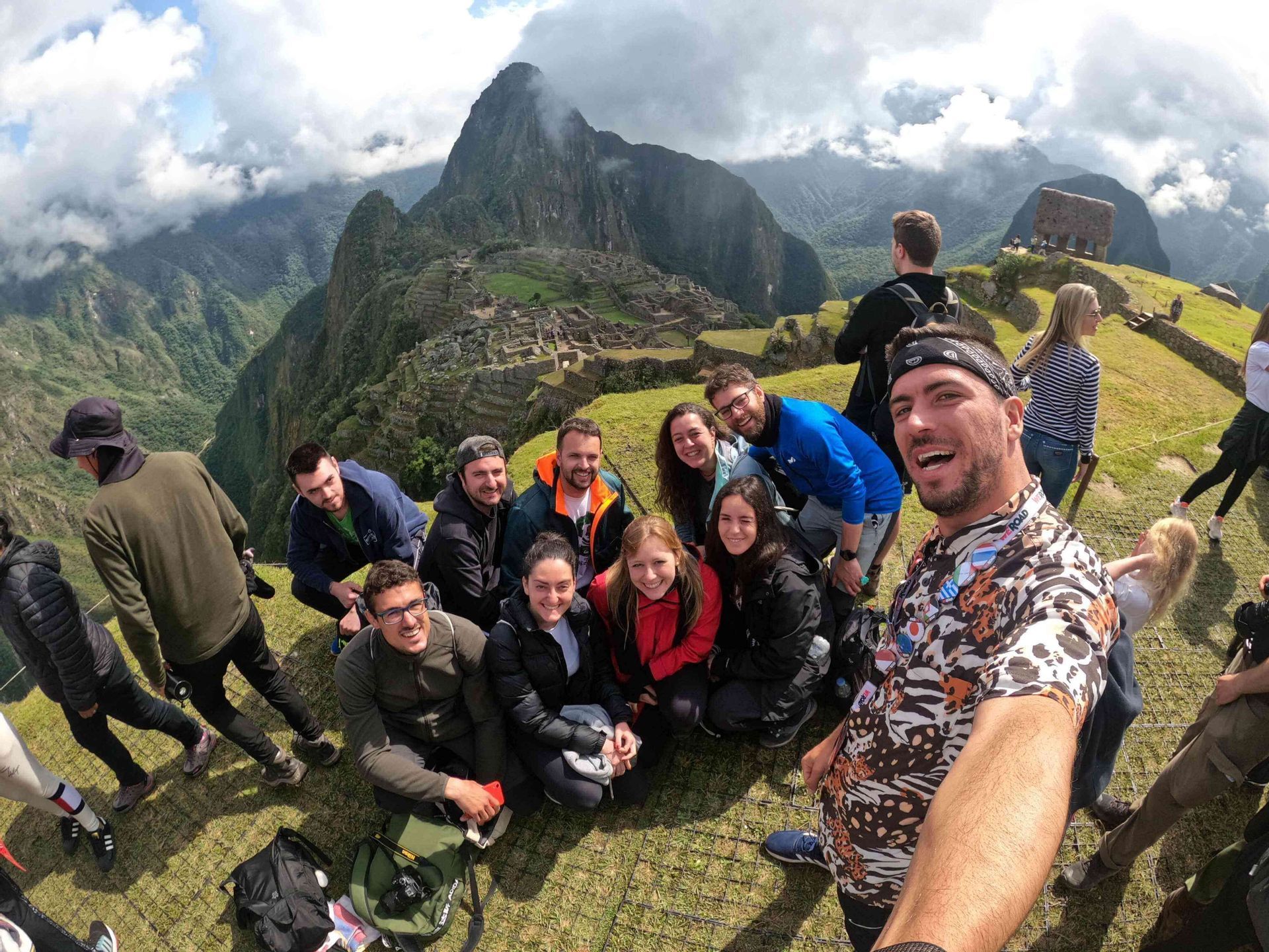 Un groupe WeRoad prend un selfie souriant sur un point de vue herbeux, surplombant des ruines antiques nichées dans des montagnes escarpées et verdoyantes.