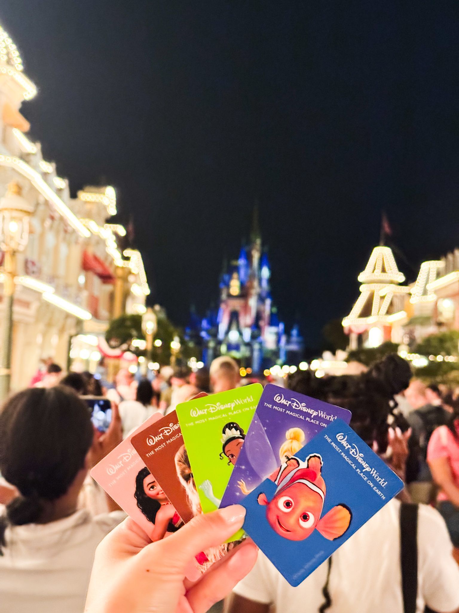 A hand holds four Walt Disney World tickets in front of an illuminated castle and crowded street at night.