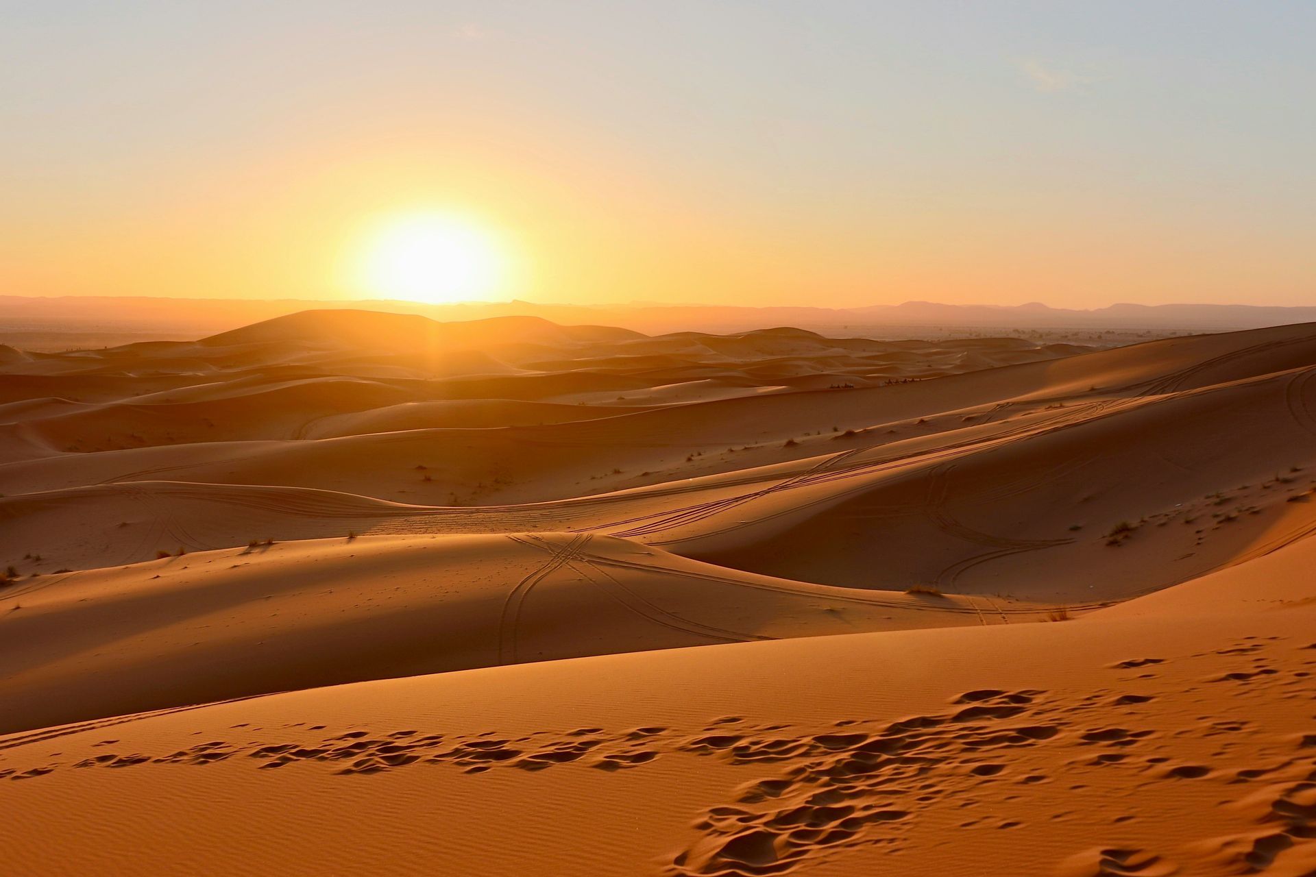 Il sole cala sulle sinuose dune di sabbia del deserto, con impronte e tracce di pneumatici visibili nella sabbia dorata.