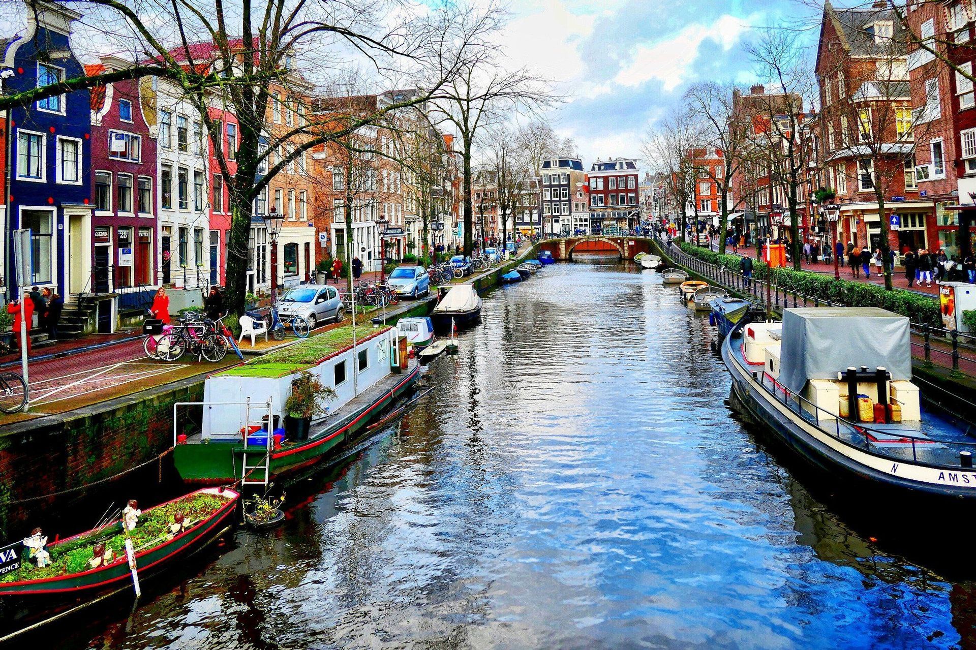 Barcos amarrados en un canal urbano bordeado por edificios coloridos, con personas caminando por la calle adyacente bajo un cielo parcialmente nublado.