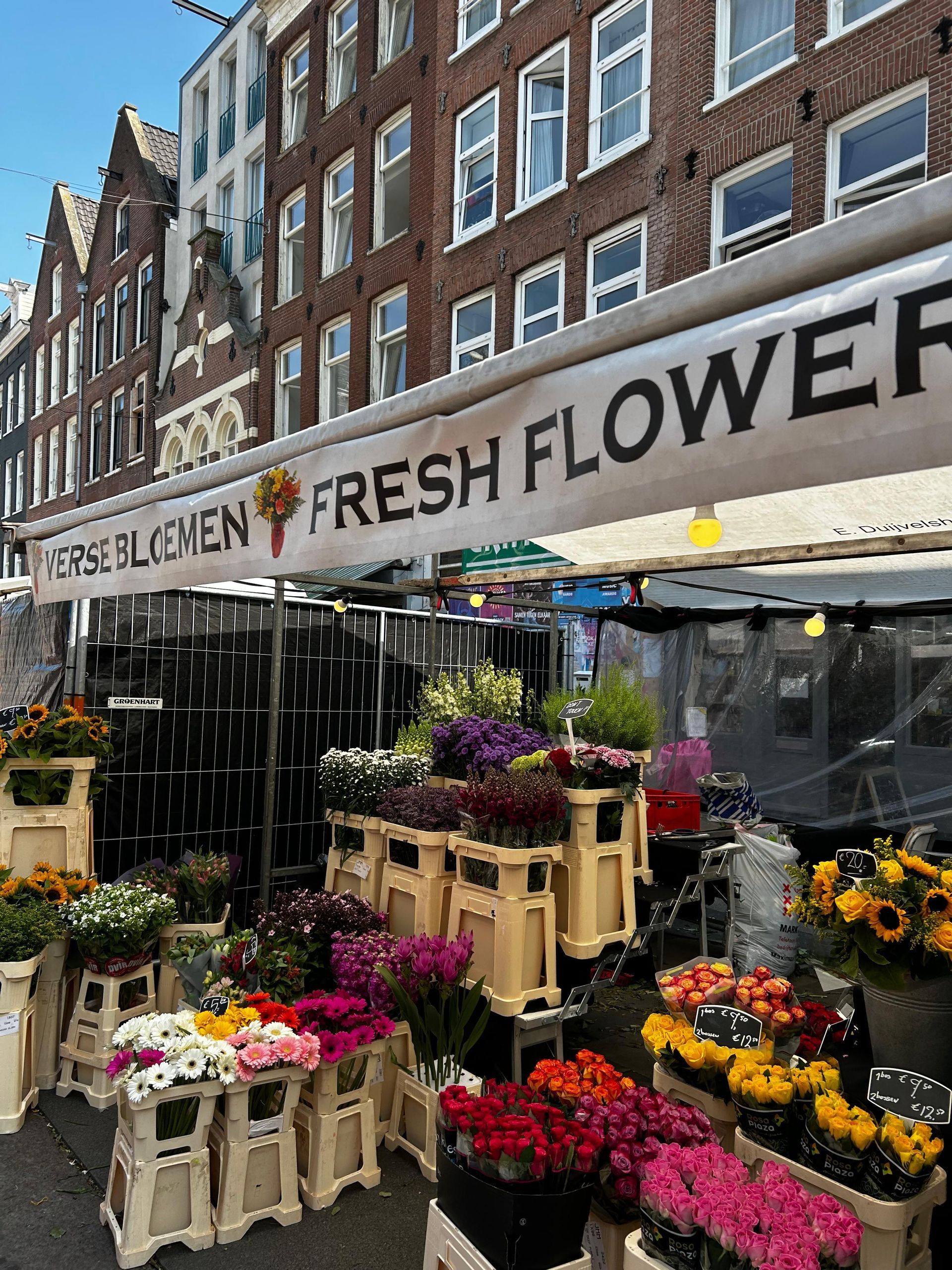 Un puesto de mercado de flores al aire libre con un cartel de 'Flores Frescas', mostrando ramos coloridos en cajas en una calle de la ciudad.