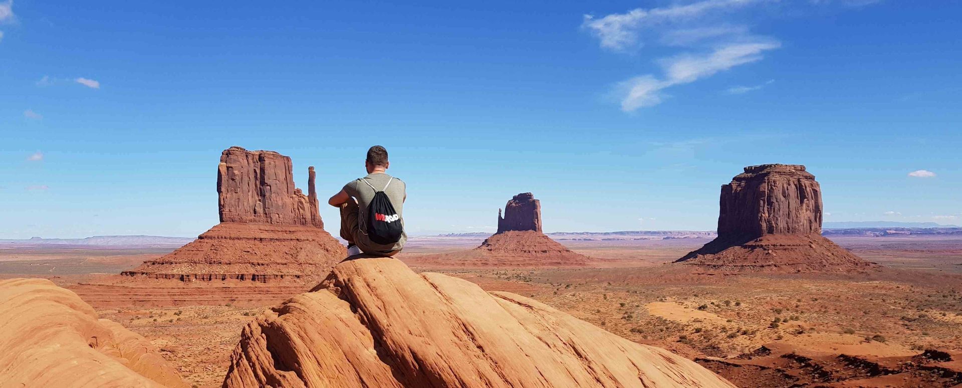 A person wearing a WeRoad backpack sits on a red rock, overlooking a desert valley with large buttes under a blue sky.