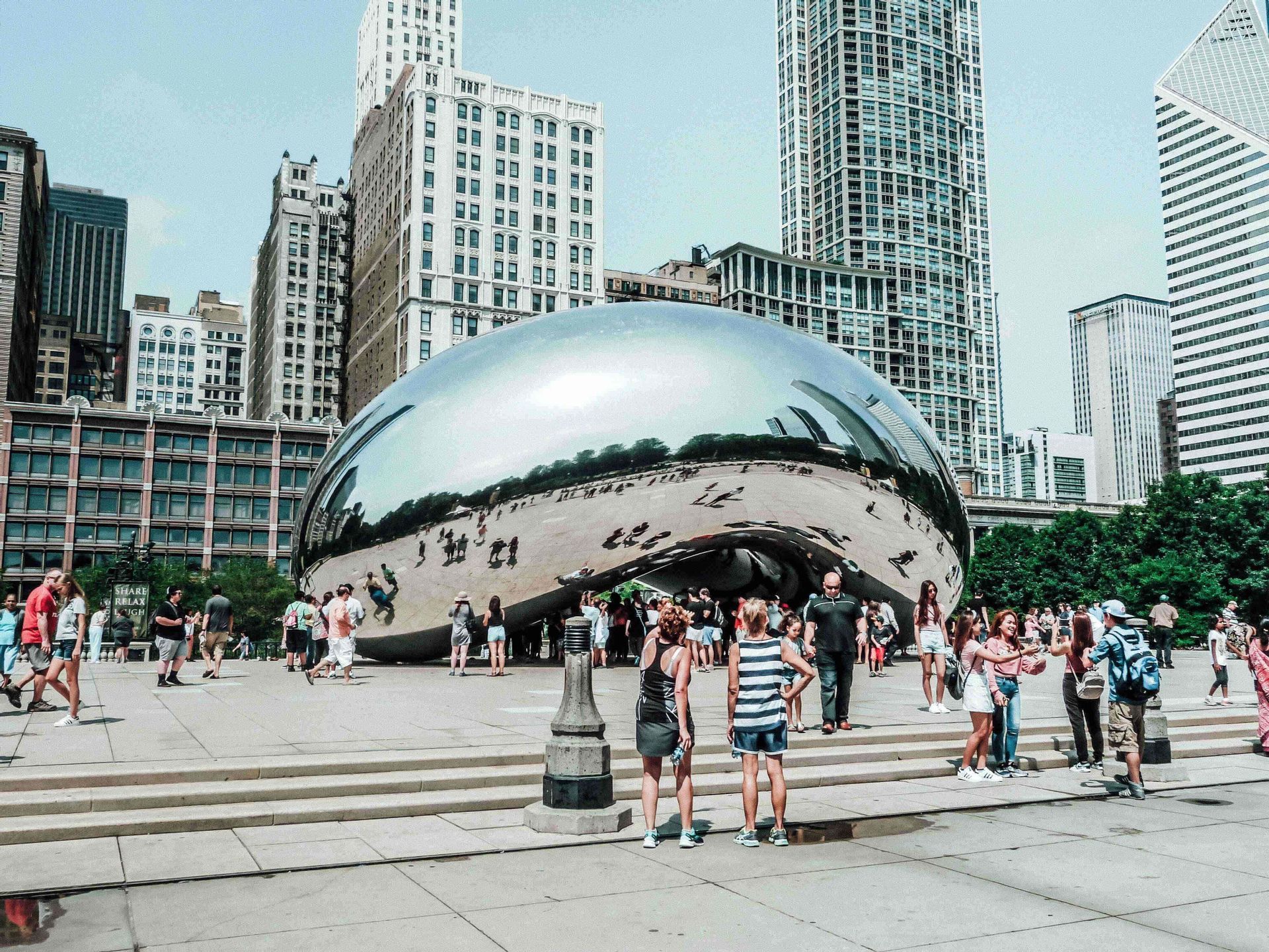 A crowd of people gathers around a large, reflective, bean-shaped sculpture in a city plaza, with skyscrapers in the background.