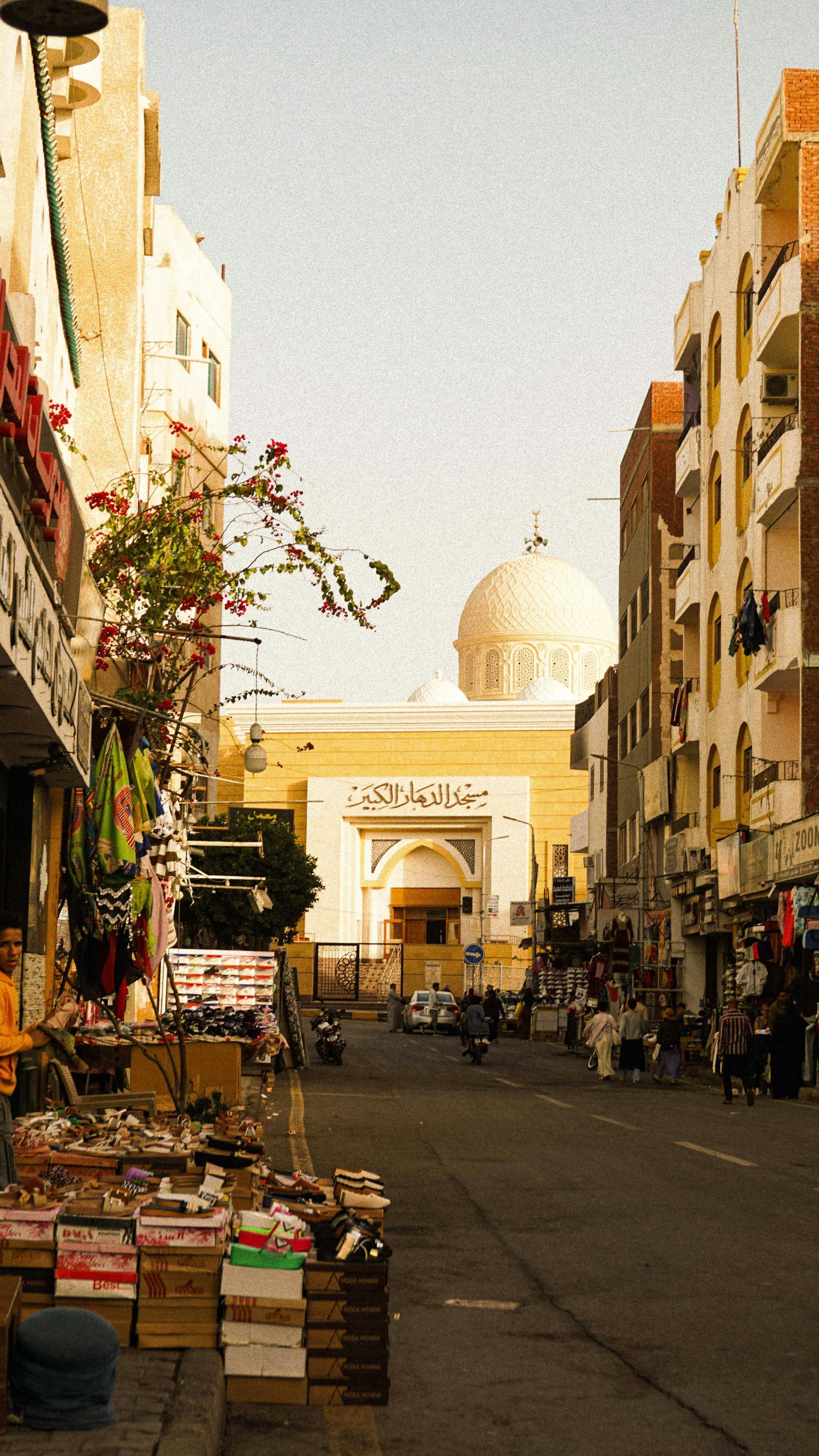 Una vista su una strada cittadina fiancheggiata da negozi, con una moschea prominente e la sua grande cupola bianca visibile sullo sfondo.