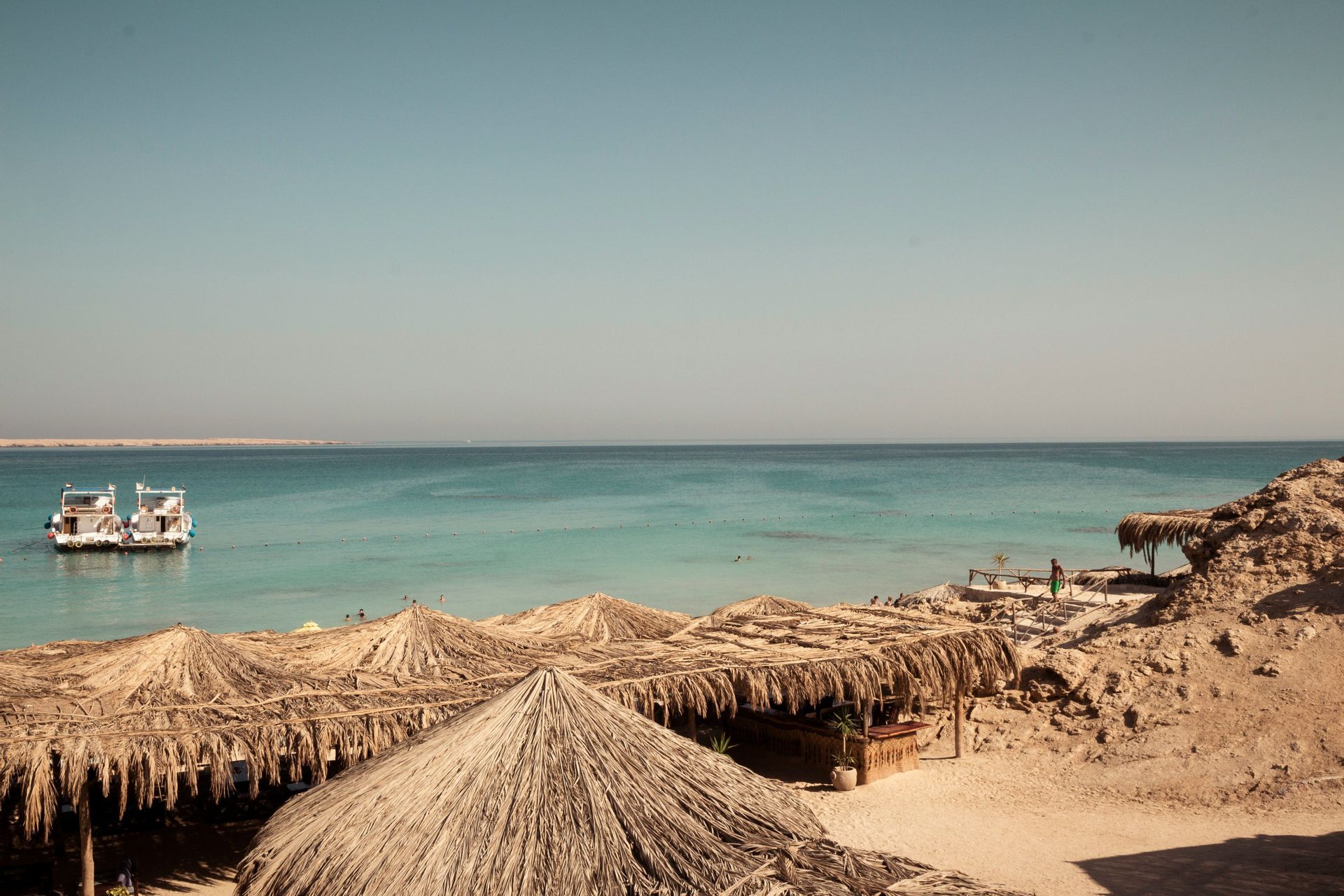 Una vista dall'alto di una spiaggia sabbiosa con ombrelloni dal tetto di paglia, che si affaccia su due barche ormeggiate in acqua turchese sotto un cielo sereno.