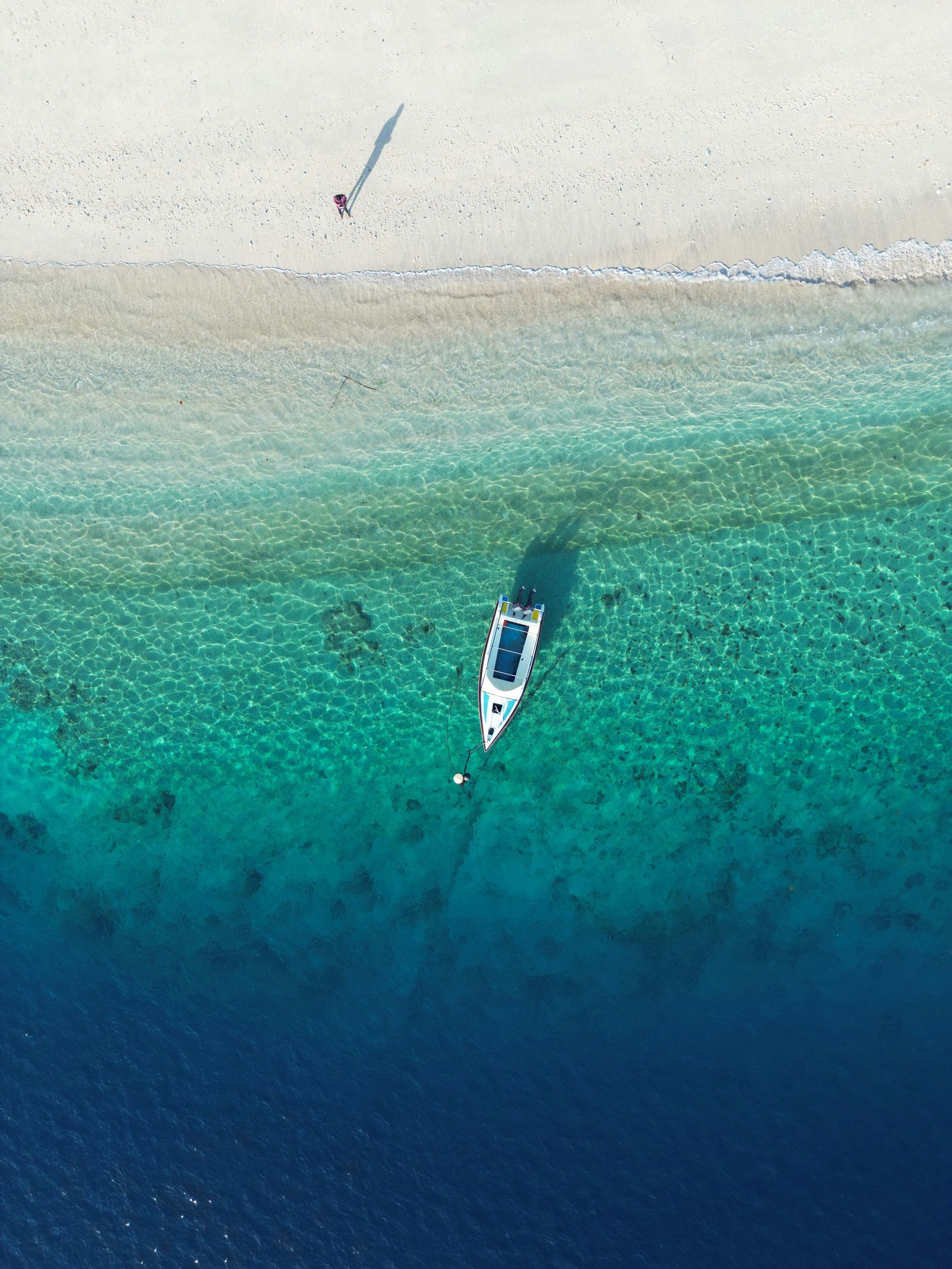 Vista aerea di una barca ancorata in acqua turchese accanto a una spiaggia di sabbia bianca con una persona che cammina lungo la riva.