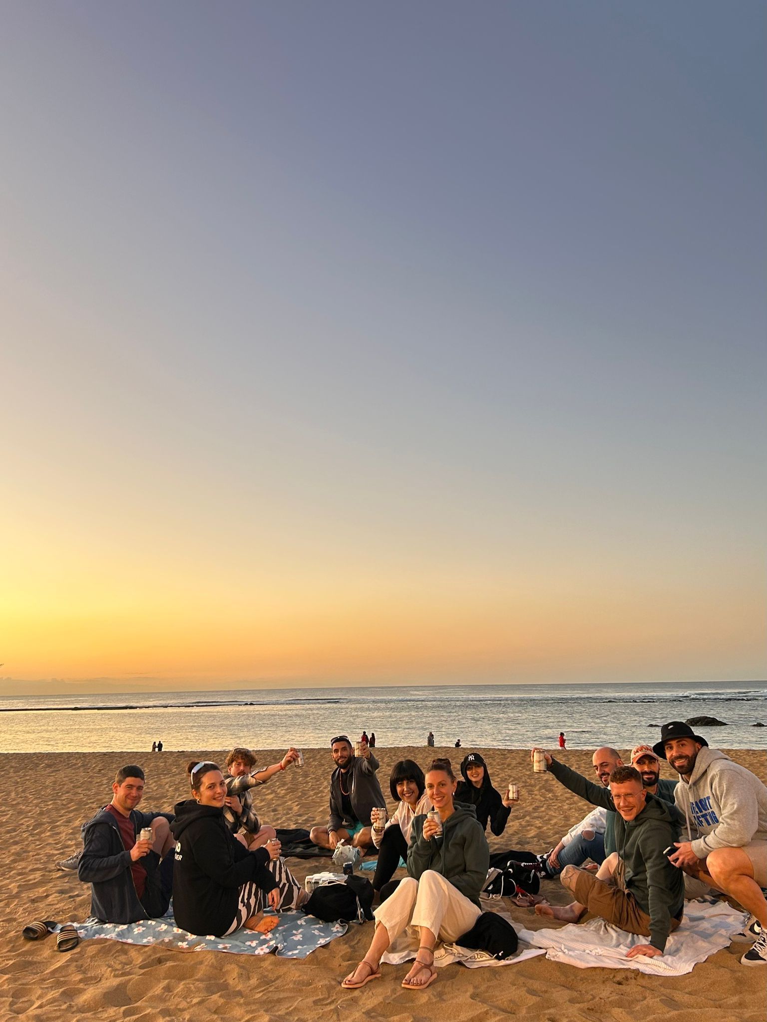 A WeRoad group trip sits together on blankets on a sandy beach, enjoying drinks during a golden sunset over the ocean.
