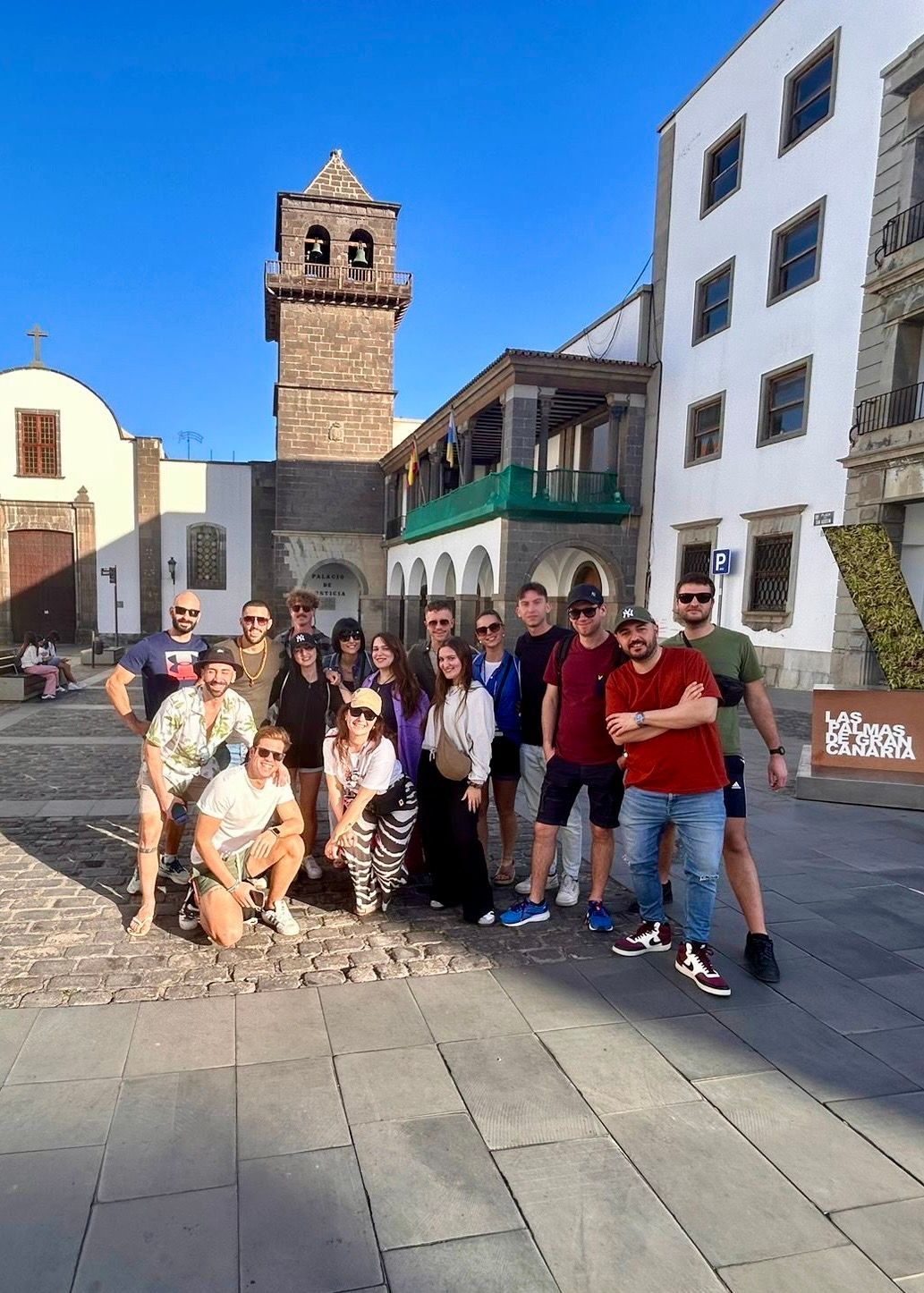 A WeRoad group trip poses for a photo in a sunny city square with a stone bell tower in the background.