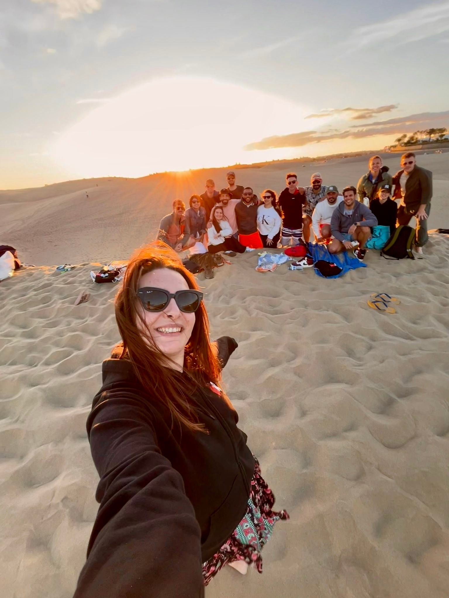 A woman takes a selfie with a WeRoad group trip as they pose together on sand dunes during a golden sunset.