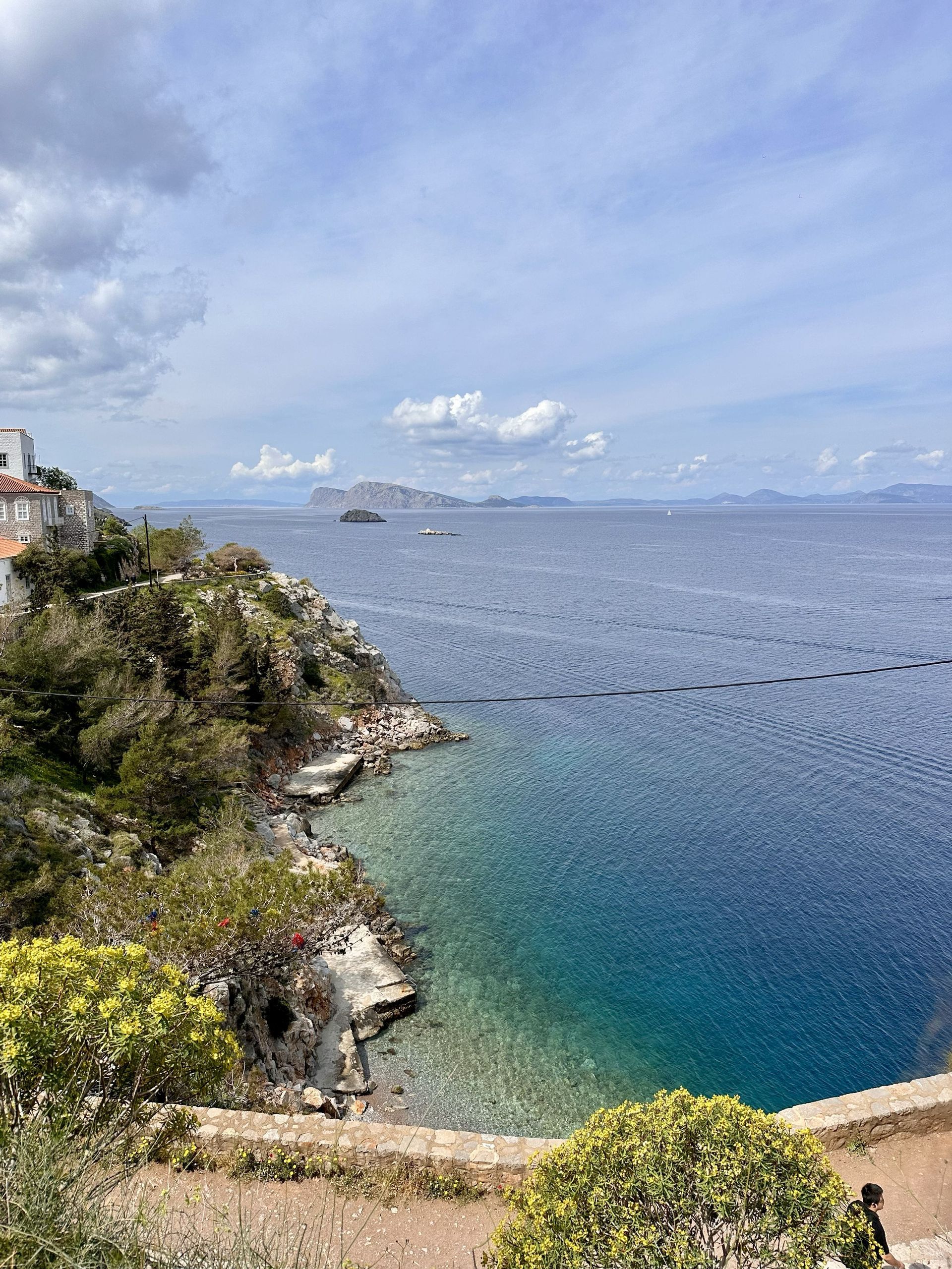Una vista dall'alto di una costa rocciosa con alberi verdi, una piccola caletta con acqua turchese e isole lontane sotto un cielo nuvoloso.