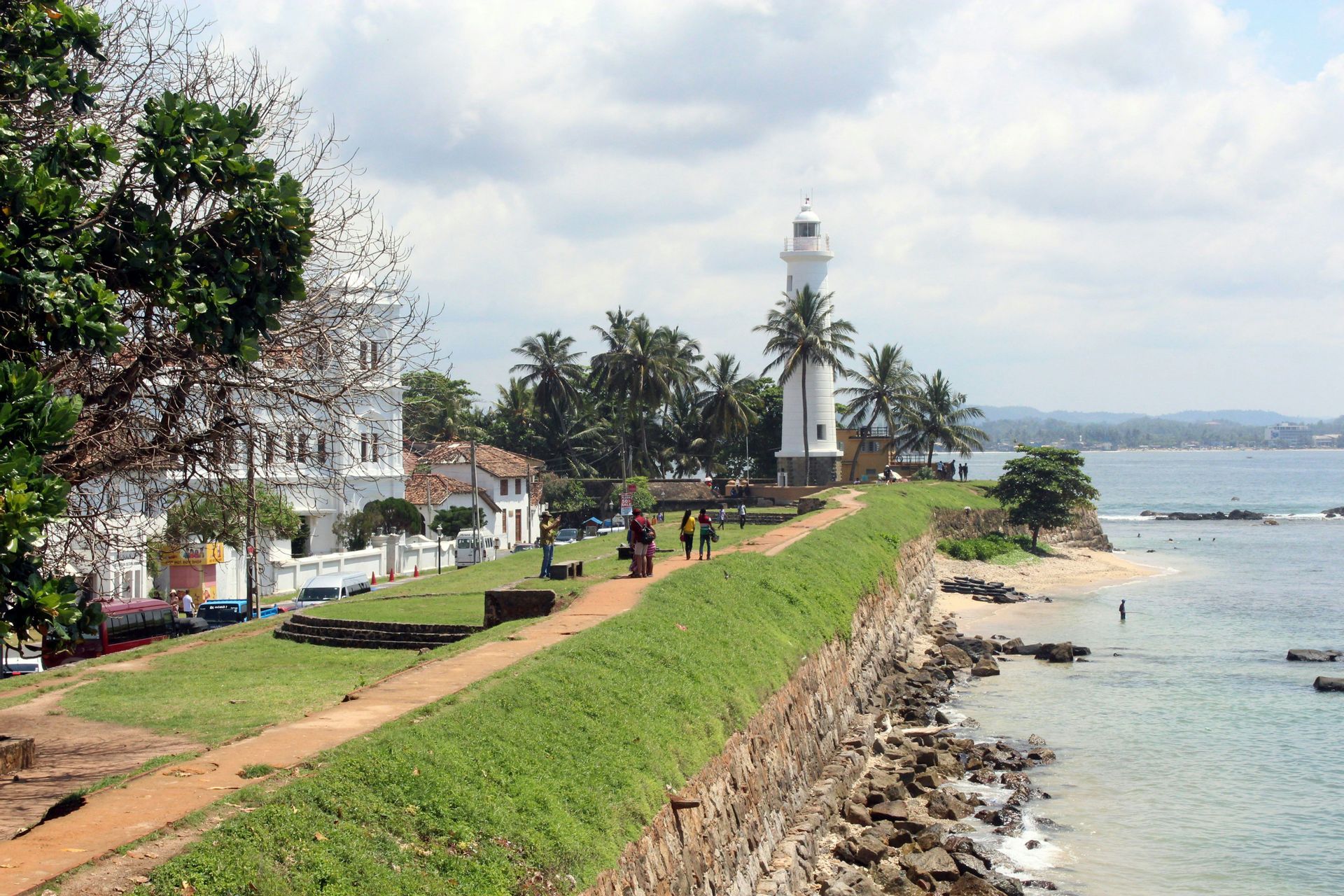 Una vista panorámica de un faro blanco y palmeras en una costa verde con un muro de piedra y el océano.