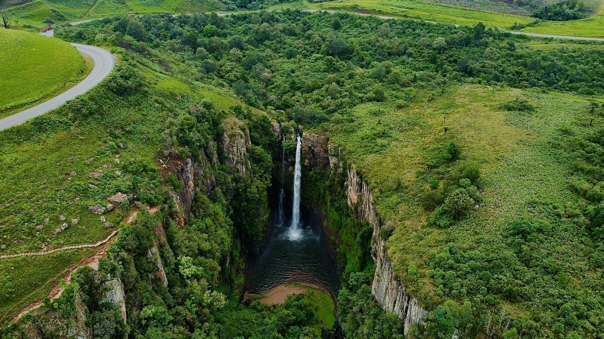 Luftaufnahme eines Wasserfalls, der von einer Klippe in ein Becken in einer üppig grünen Schlucht stürzt, umgeben von sanften Hügeln.
