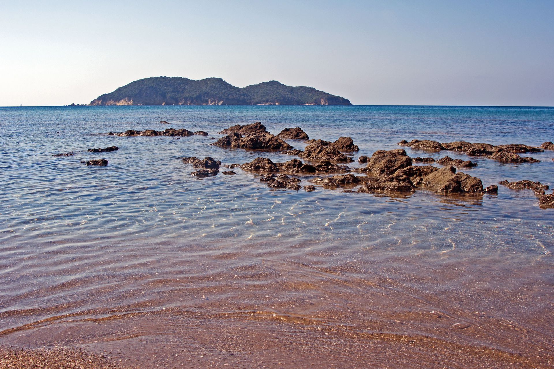 Ein felsiges Ufer mit klarem, flachem Wasser, das an einen Sandstrand schwappt, mit einer grünen Insel am Horizont.
