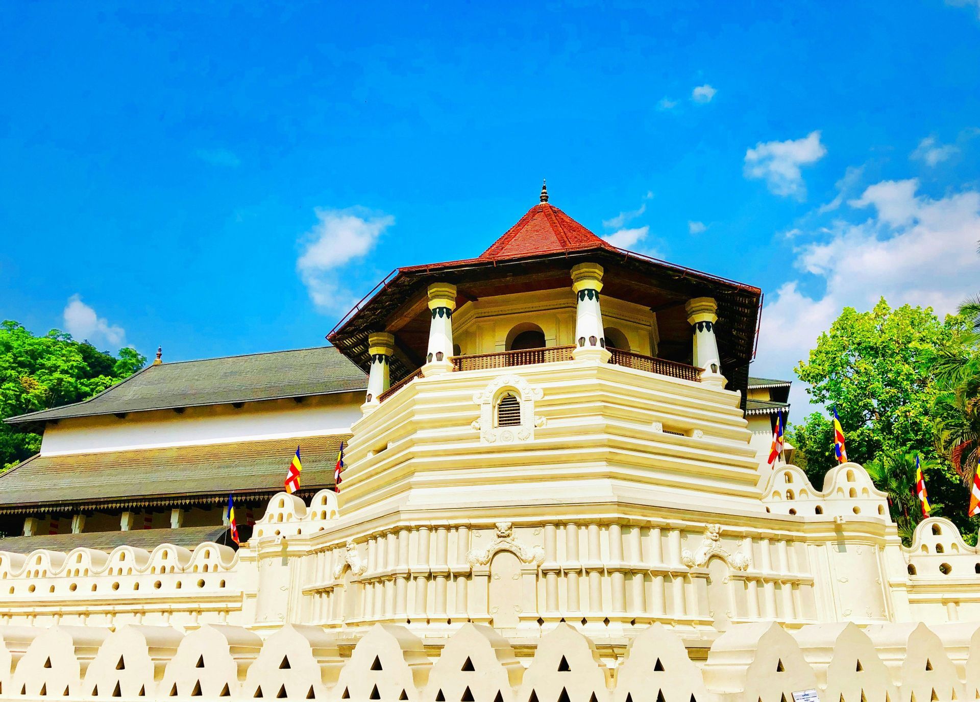 Ein mehrstufiger weißer Tempel mit einem achteckigen Turm und rotem Dach steht hinter einer dekorativen Mauer unter blauem Himmel.