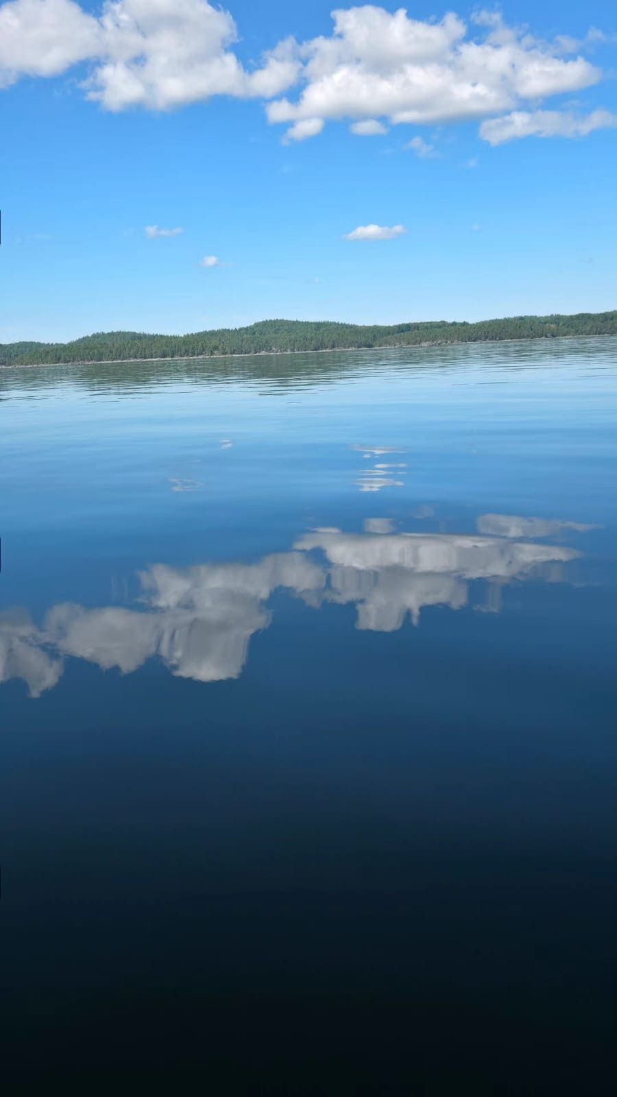 Ein ruhiger See spiegelt den blauen Himmel und die weißen Wolken darüber perfekt wider, mit einem bewaldeten Ufer im Hintergrund.