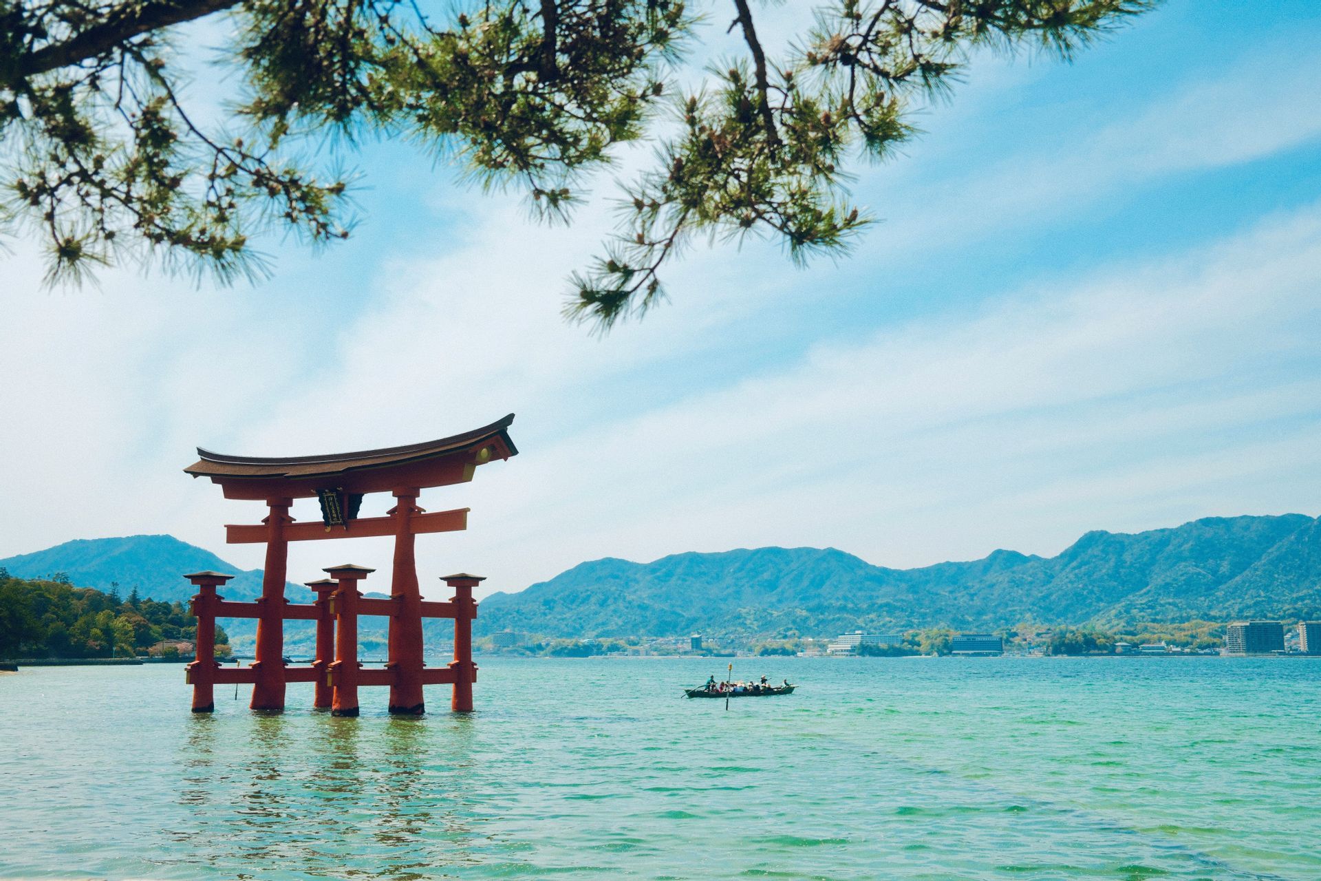 Ein rotes japanisches Torii-Tor steht im Wasser, umrahmt von einem Kiefernast, mit Bergen und einem Boot im Hintergrund.