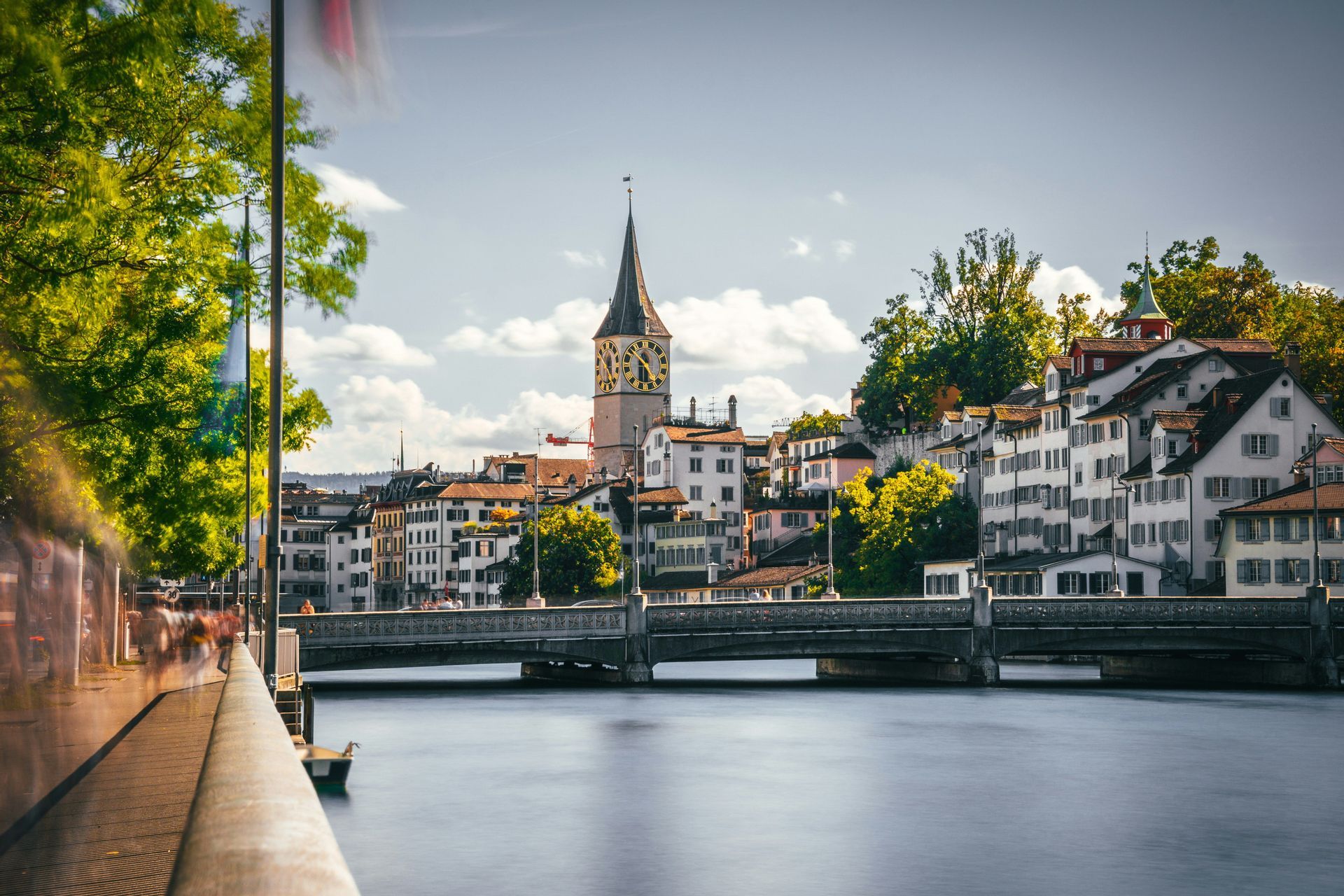 Eine Stadtansicht mit markantem Uhrturm und einer Flussbrücke, von einer baumgesäumten Promenade aus gesehen.