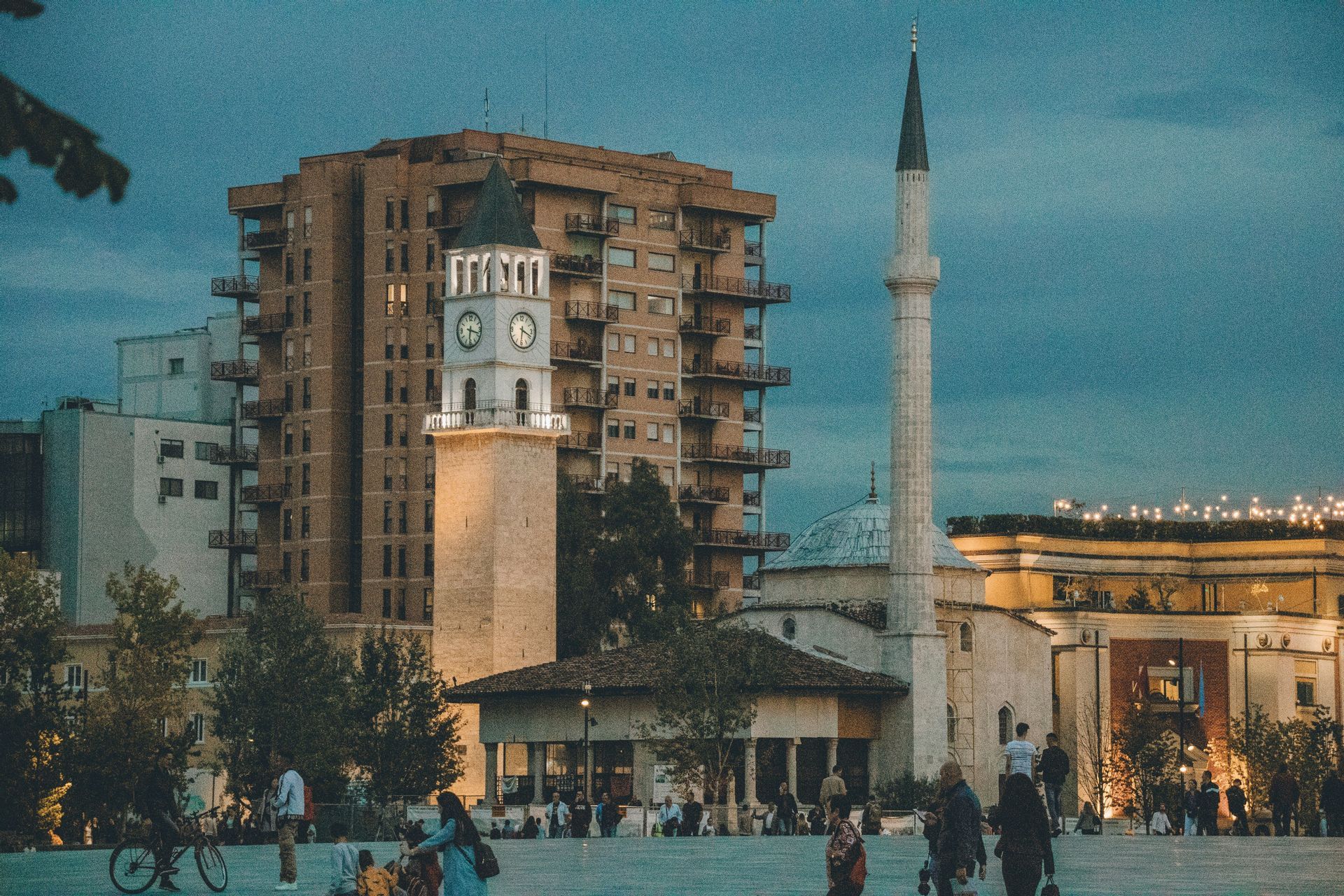 An illuminated clock tower and a mosque's minaret stand in a city square at dusk, with people walking in the foreground.